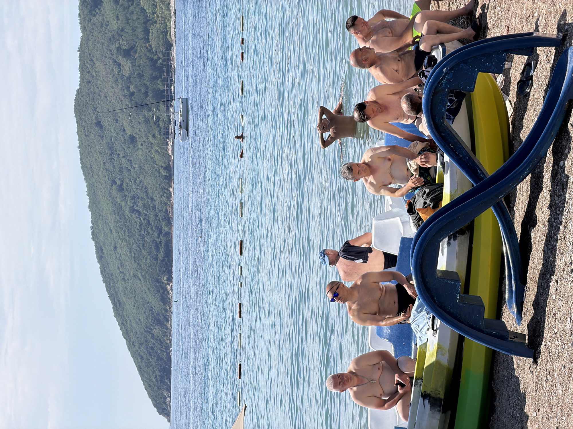 This horizontally oriented image captures a sunny beach scene in Budva, Montenegro. The foreground and midground are dominated by a group of approximately eight to ten adult men, mostly middle-aged or older, relaxing on and around paddle boats pulled onto a pebbly beach. Most of the men are shirtless, wearing swim trunks, and appear to be enjoying a leisurely day by the sea.
Several pedalos, including blue and white, and yellow and green models, are visible on the shore; one features a blue slide attachment. Some men are seated on these boats, while others are on the sand. Notable details among the individuals include one man in the foreground wearing a cross necklace and another in the midground wearing sunglasses.
The calm, blue sea stretches across the midground, marked by a line of buoys. One person is partially submerged in the water, possibly swimming or wading. In the background, a large, densely forested mountain rises from the coastline, and a sailboat is visible further out at sea. The sky is clear with a few wispy clouds, and strong shadows indicate bright daylight, suggesting it is mid-morning or afternoon. The overall scene conveys a relaxed, vacation-like atmosphere in a beautiful coastal setting. No visible text or discernable logos are present.