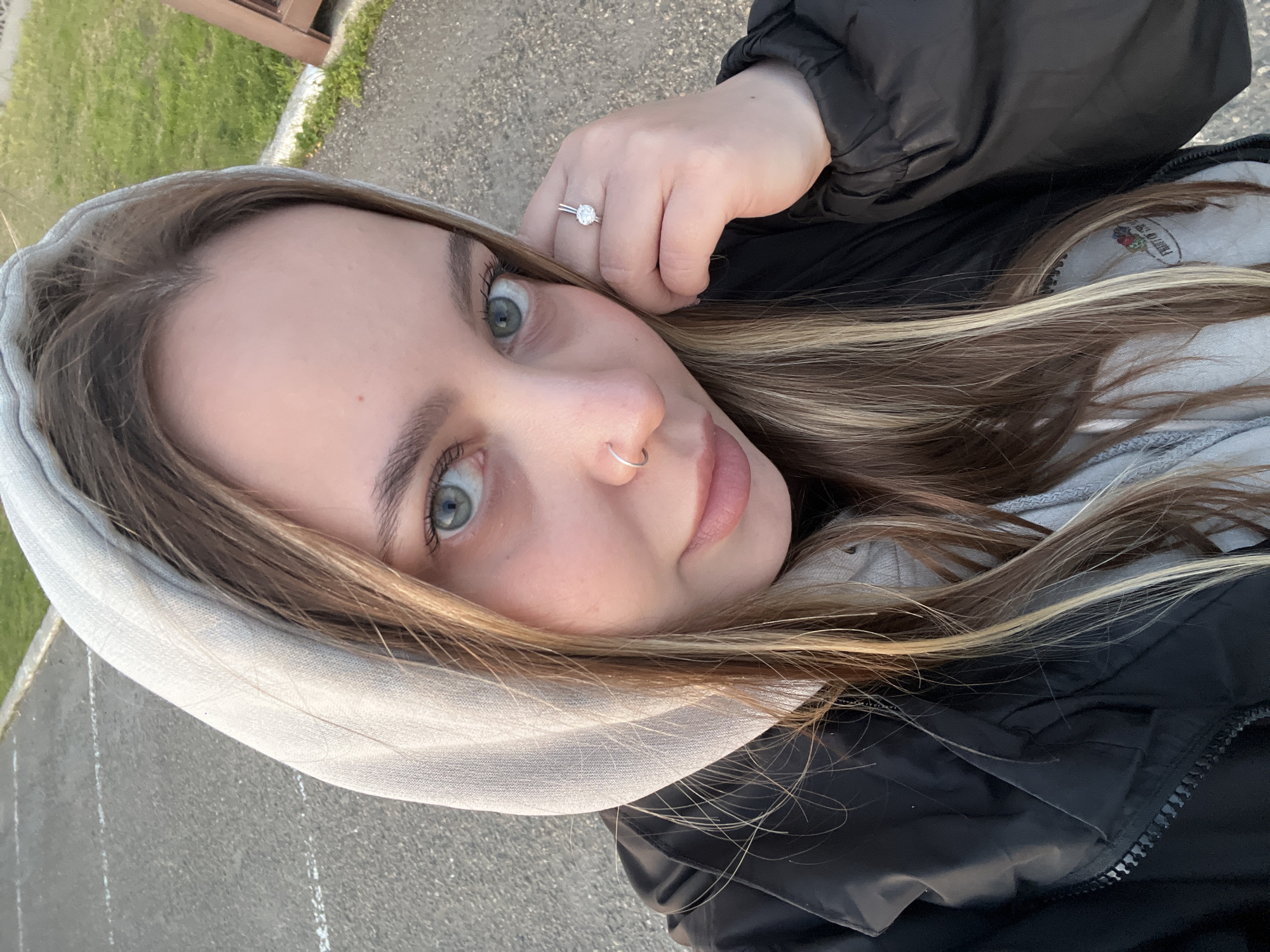 A close-up, angled selfie captures a young woman outdoors in Novopokrovskaya, Russia, during daytime. The woman, likely in her late teens or early twenties, has long light brown hair with prominent blonde highlights. Her striking blue-green eyes gaze directly at the camera, and she has a silver hoop nose ring on her right nostril. On her left ring finger, a silver ring with a clear, round stone is visible as her hand is raised near her face.
She is dressed in casual, warm attire, wearing a light grey hooded sweatshirt underneath a black puffer jacket. A faint logo, identifiable as "Fruit of the Loom" with its characteristic fruit cluster design (apple, grape, and leaves), is visible on the grey hoodie near her right shoulder. The background features a paved surface, possibly a road or pathway, with some white markings. Green grass and a distant brown or wooden structure are also visible, suggesting an outdoor setting like a park or roadside. The lighting is soft and natural, indicating daylight, with no harsh shadows. Her expression is calm and direct.