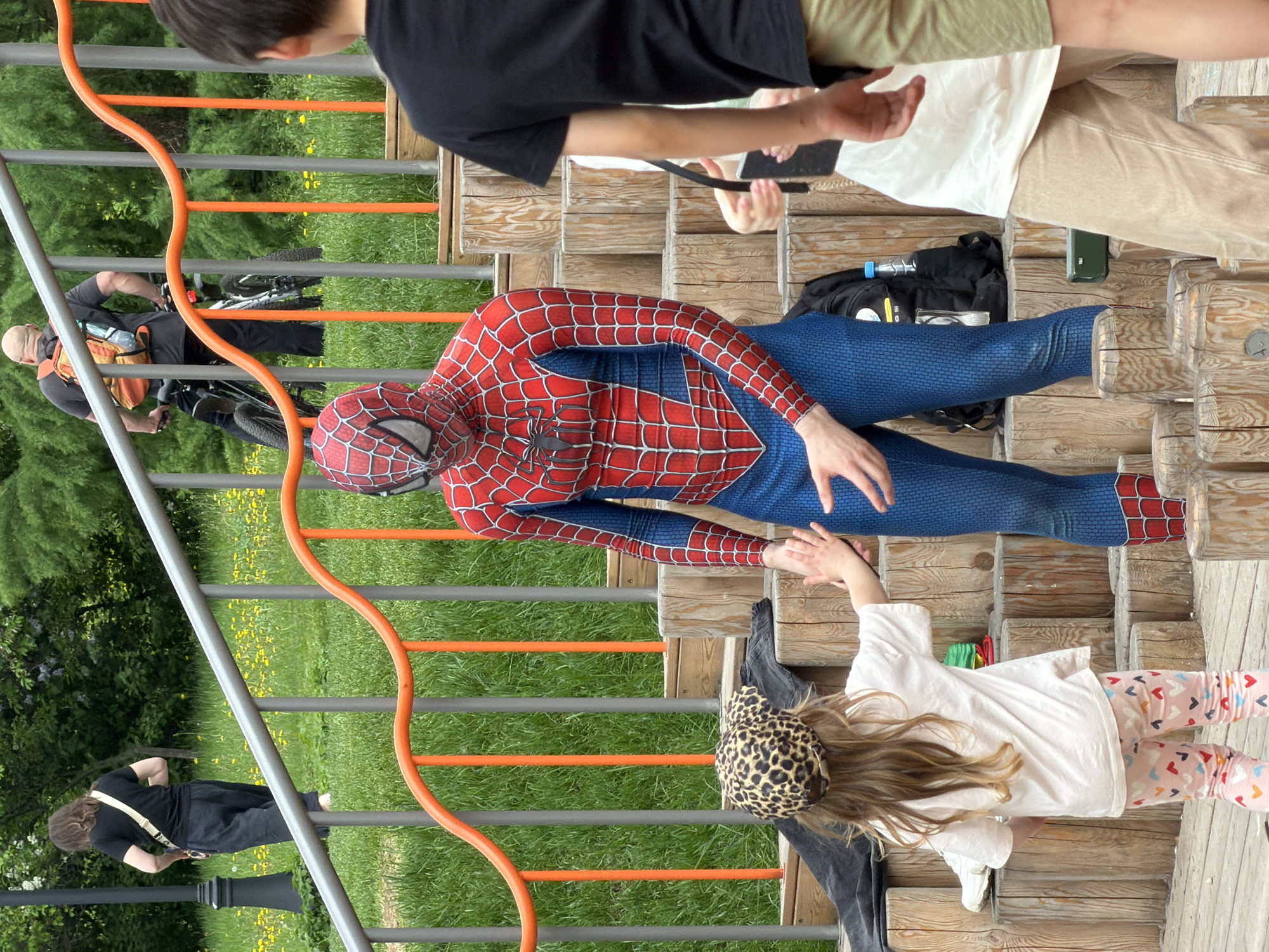 The image shows a person dressed as Spider-Man interacting with a child on a wooden structure that resembles a playground or staircase.
People:
- A person in a Spider-Man costume, posed to interact with the child.
- A young child reaching towards Spider-Man. The child is wearing a white shirt, leggings with heart patterns, and a leopard-print headband.
- Other people are present in the background, including individuals walking and one person with a bicycle.
Objects:
- A wooden structure, possibly part of a playground or set of stairs, made of log-like wooden steps.
- Metal railings with orange horizontal bars adjacent to the wooden structure.
- A backpack, a water bottle, and a phone placed near the structure.
- Bicycles in the background.
Actions and Interactions:
- The central interaction is Spider-Man reaching out to the child.
- Other people are walking in the area, suggesting it's a public space.
Environment:
- The scene is outdoors, likely a park or recreational area with grass and some trees.
Location:
- Moscow, Russia: This location is implied by the identification tag.