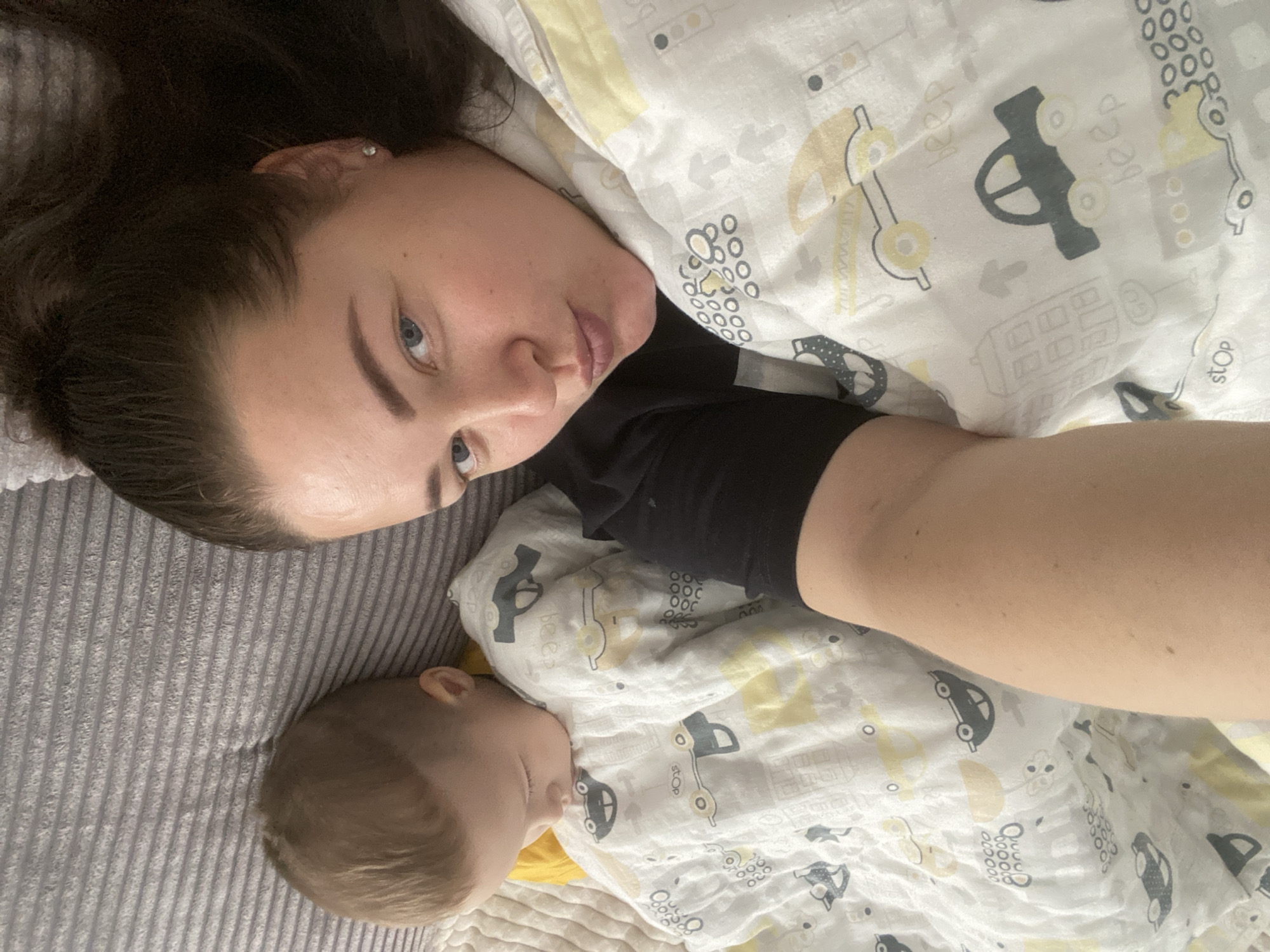 The image captures a person with brown hair and blue eyes lying next to a baby. The person's face is angled toward the camera. The baby is sleeping, wrapped in a blanket with a vehicle pattern. The setting appears to be indoors, likely a bedroom, with a gray ribbed blanket under the subjects and a patterned white blanket covering them. The location is not discernible as being in the city of Sochi, Russia from the given visual information.
