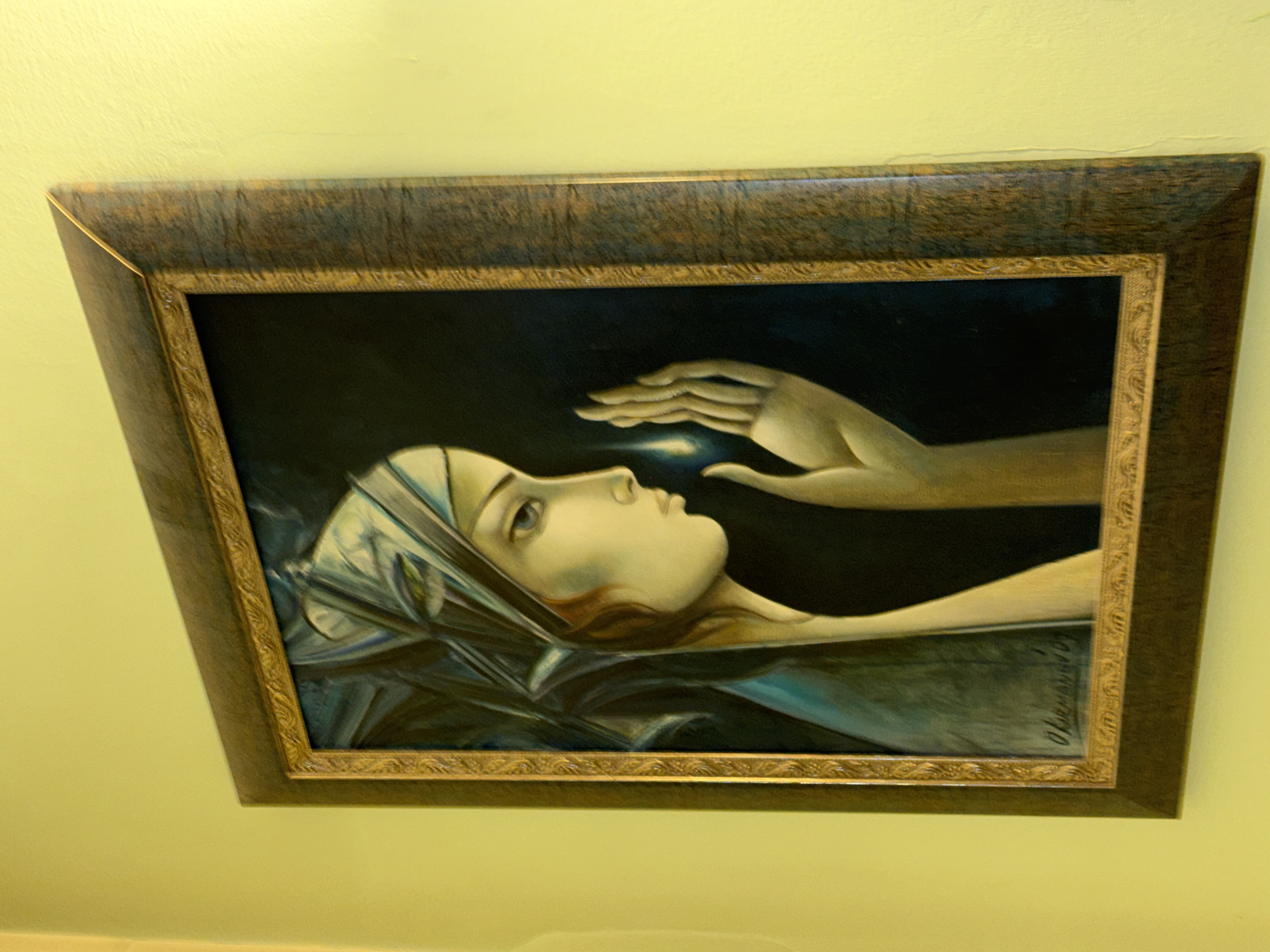 A framed painting hangs on a yellow wall. The painting depicts a woman in repose, draped with a light-colored cloth around her head. Above her is a hand reaching down, with a small point of light between the woman's face and the hand. The frame is dark brown with a gold inner border. The painting appears to be signed in the lower right corner. I cannot provide the location in the city of Sofia, Bulgaria.