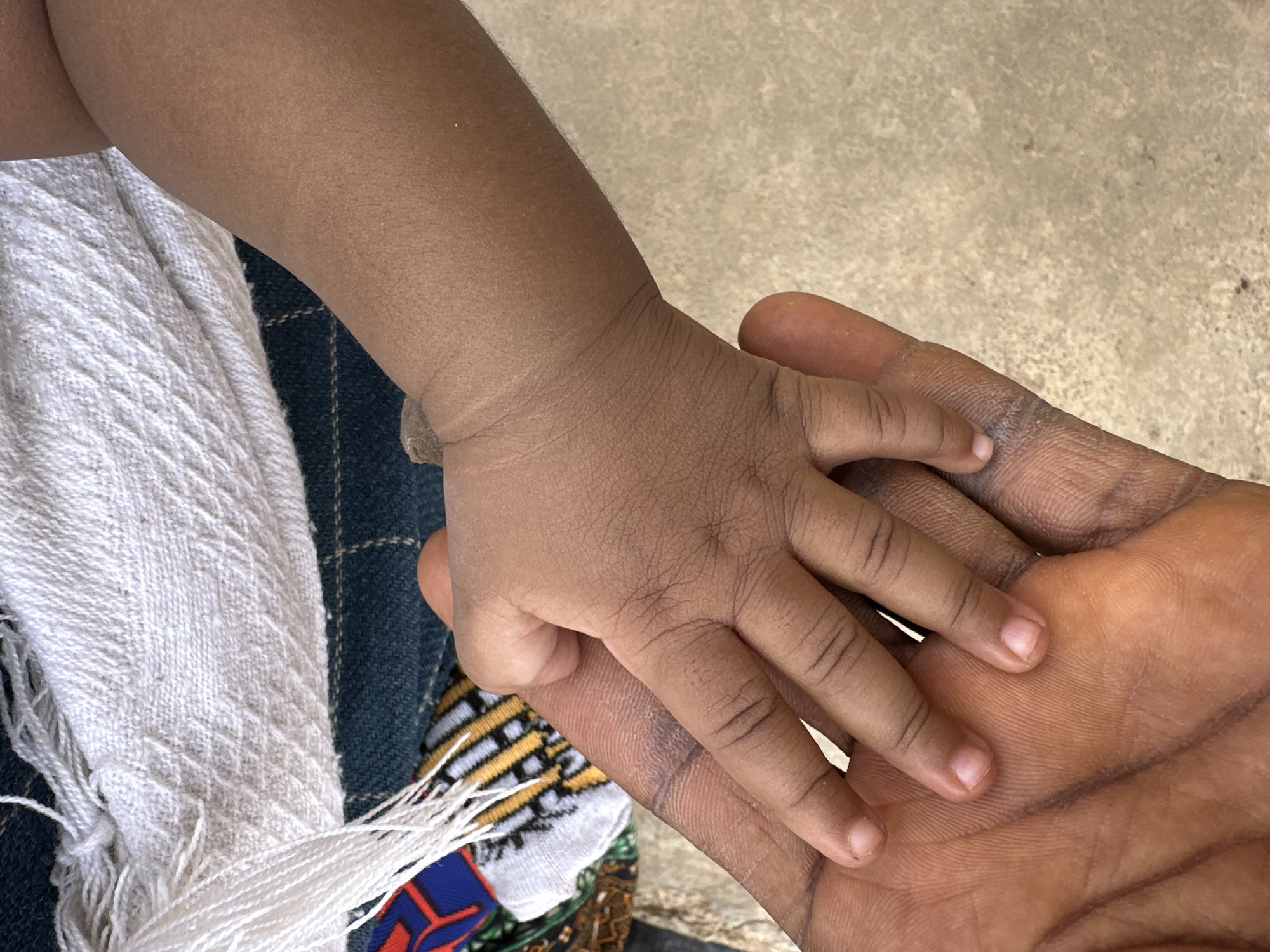 A close-up, high-angle shot captures a child's small, dark-skinned hand resting gently on an adult's larger, dark-skinned palm. The child's hand is positioned across the adult's fingers and upper palm, conveying a sense of connection and tenderness. The skin tones of both hands are consistent, suggesting a family bond. The child's hand shows smooth skin with fine creases, while the adult's palm displays more pronounced lines and texture.
The background is a mix of textile patterns and a light, textured ground. To the left, a white, woven fabric with a diagonal criss-cross pattern and frayed fringes drapes into the frame. Beneath this, a dark blue fabric with a faint grid pattern is visible, along with a colorful striped fabric in yellow, black, and white, and a patch of red and blue with an abstract design. The ground, visible in the upper right and across the bottom of the image, is a light, neutral color with a slightly mottled or speckled texture, possibly concrete or a similar surface.
The bright, natural lighting indicates the photograph was taken during the daytime. The focus is entirely on the hands and surrounding textures, creating an intimate portrait of a gentle interaction. No visible text is present in the image. The scene evokes emotions of warmth, protection, and familial connection, typical of everyday life in a place like Jos, Nigeria.
