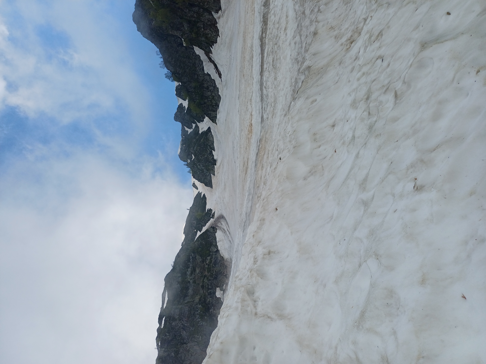 The media file depicts a snow-covered mountainside with a rocky outcrop. The scene is dominated by the stark contrast between the dark, rugged rock and the white, textured snow. Patches of green vegetation are visible clinging to the rock face, suggesting a mountain environment. The sky above is a light blue, with scattered white clouds, indicating fair weather.
The perspective is from a low angle, looking up at the slope. The snow appears somewhat uneven and textured, with darker streaks and footprints or irregularities suggesting recent melting or disturbance. The rocky formation is steep and appears to be a cliff face or a steep slope.
There are no people or objects clearly visible in the scene. The activity appears to be a natural landscape, possibly in a mountainous region during a season where snow is still present but melting. The lighting suggests it could be daytime, perhaps late morning or early afternoon, given the brightness of the sky and the illumination of the snow.
The location cues point towards a mountainous region like Estosadok, Russia, known for its alpine landscapes. The overall impression is one of a rugged, natural, and somewhat imposing mountain environment. No text is visible in the media file.