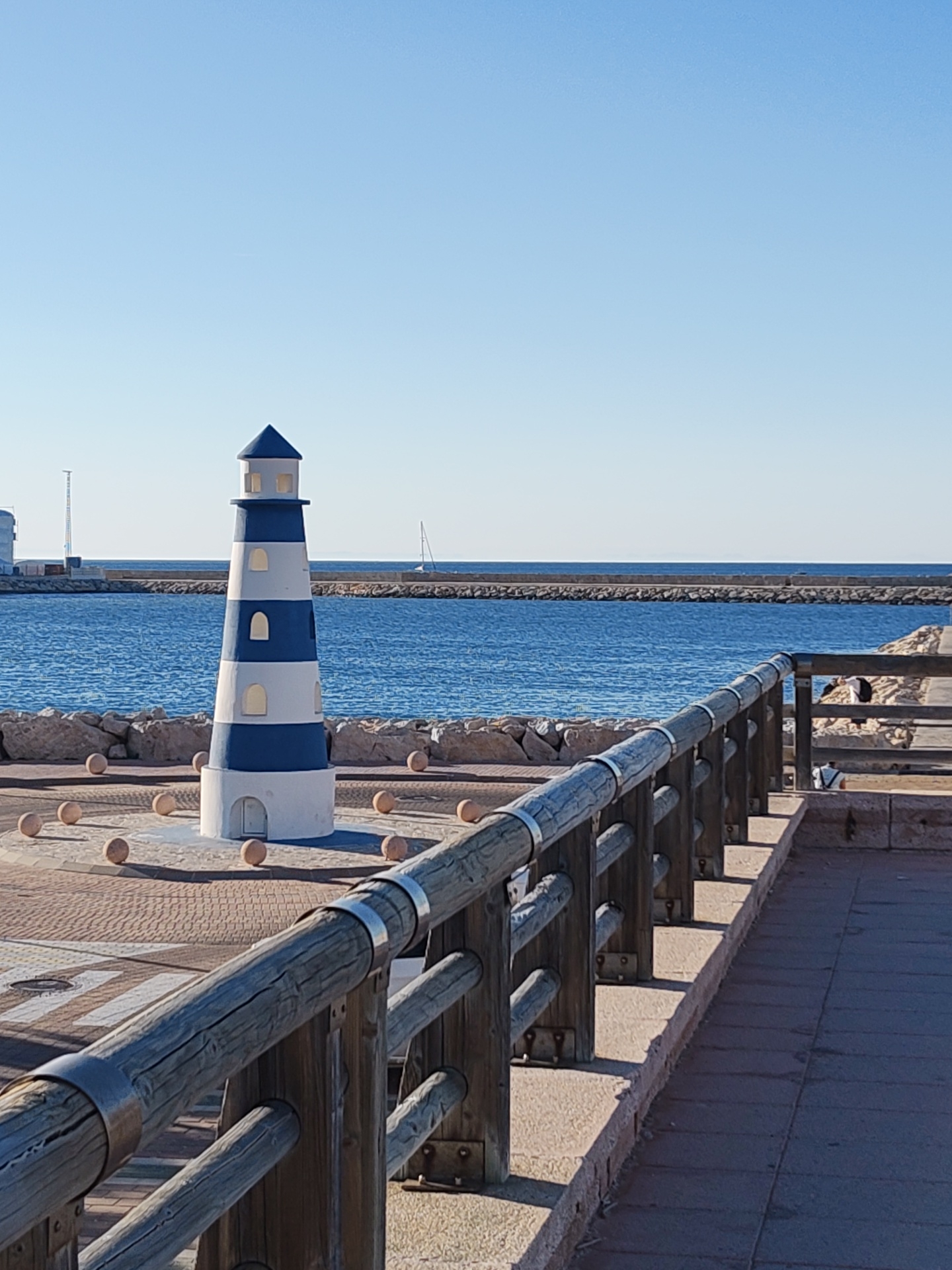 The image shows a maritime scene in Dénia, Spain, featuring a decorative lighthouse model on a paved promenade. The lighthouse is painted with alternating blue and white horizontal stripes. It stands on a circular base surrounded by spherical stones. In the background is a blue sea, bordered by a breakwater. A single sailboat is visible on the horizon. The right foreground features a wooden railing. People are partially visible in the background beyond the railing. The sky is clear blue.