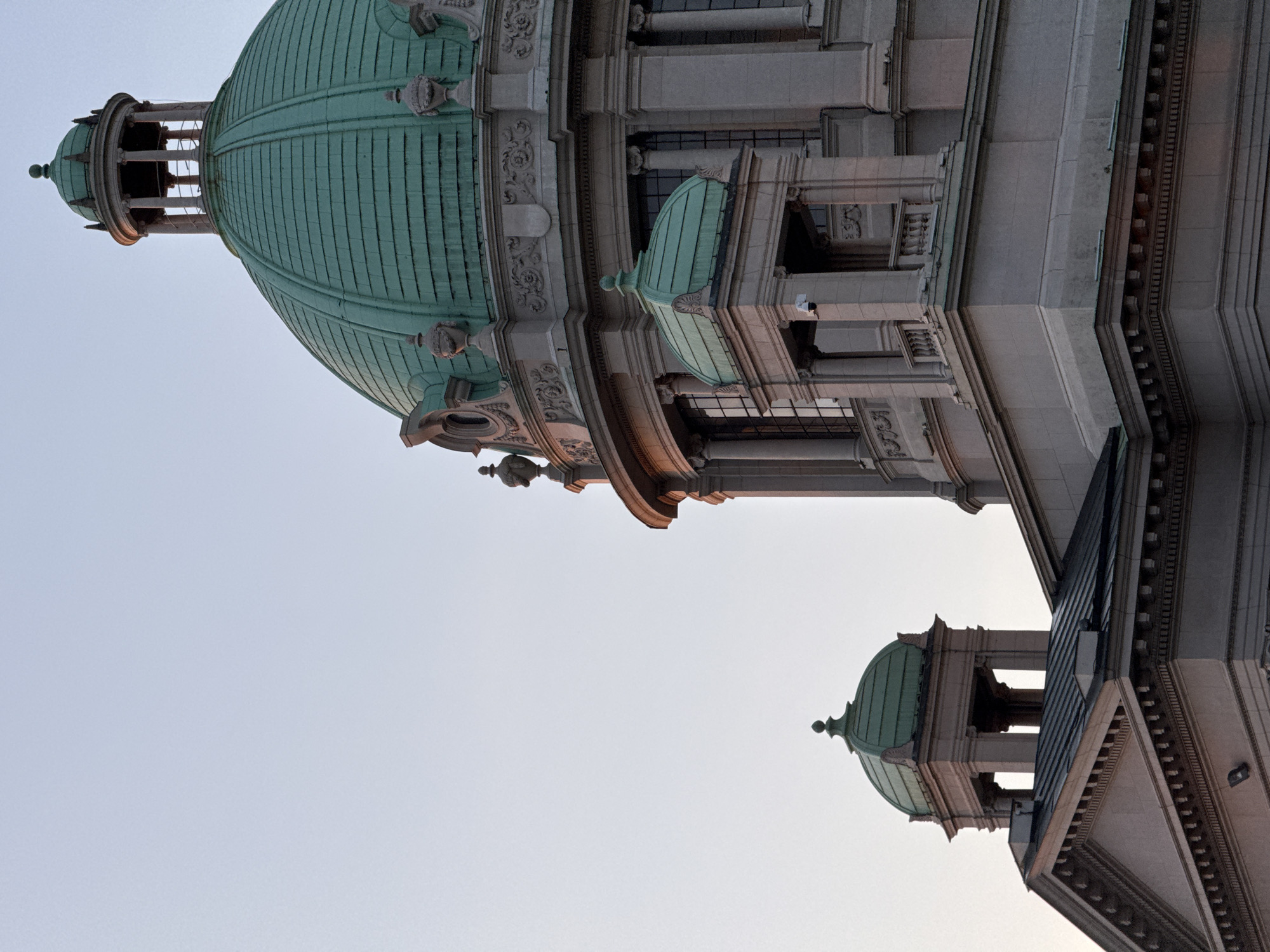 The image shows a close-up of the dome and a smaller turret of the National Assembly building in Belgrade, Serbia, at sunset. The architecture is neoclassical, with a large, green copper dome and a smaller, green copper-topped turret. The building is made of light-colored stone, with ornate carvings and columns visible. The sky is clear and pale blue, with a hint of orange from the setting sun. The image is a detailed architectural study, highlighting the building's grandeur and historical significance.
