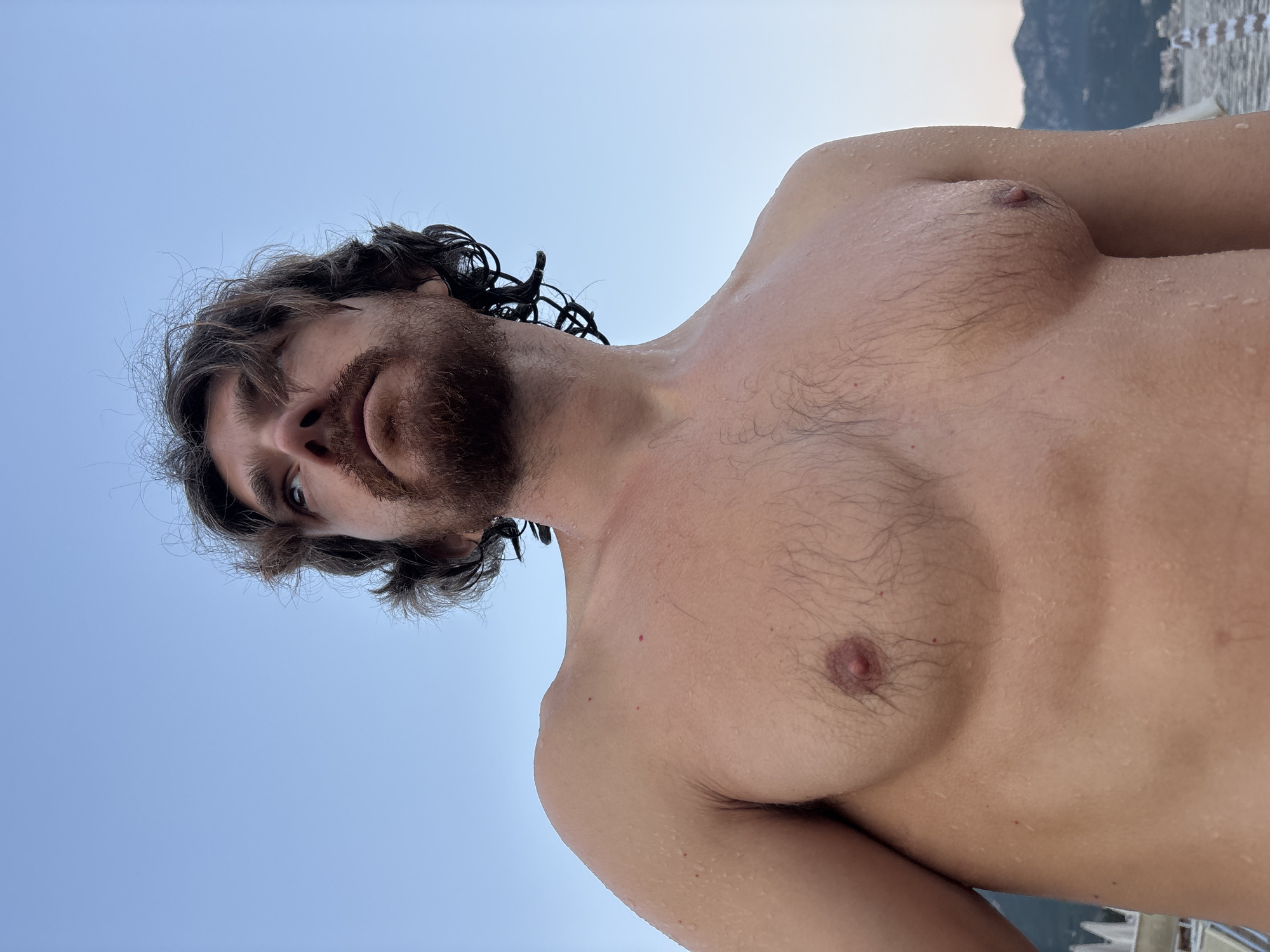 The image shows a close-up, low-angle shot of a shirtless man with wet, curly brown hair and a beard, looking upwards against a clear sky. He is positioned on a beach, with the sea and a mountainous coastline visible in the blurred background. The man's chest and shoulders are visible, with water droplets on his skin. In the far background, a white structure with a row of upright elements can be seen, possibly part of a beach resort or pier. The overall impression is one of being outdoors in a coastal area.