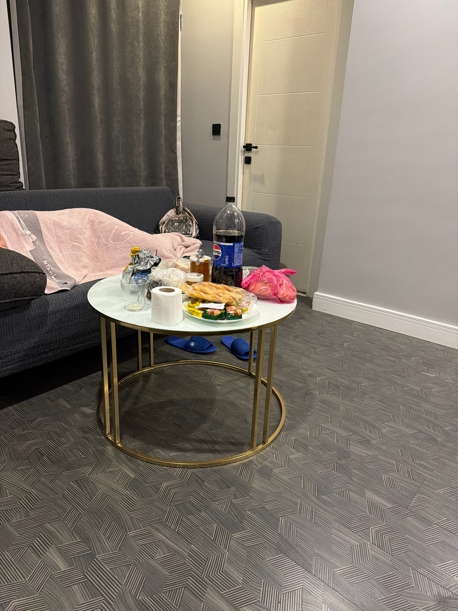 The image depicts a living room area, likely in an apartment in Ataşehir, Turkey. A dark grey sectional sofa is partially visible on the left, covered with a pink blanket. A round, white-topped coffee table with a gold metal frame stands in the foreground. On the table are various items, including a roll of toilet paper, a glass with a drink, snacks in bags and on a plate, a jar of honey, and a large bottle of Pepsi. A pink plastic bag is also on the table. A pair of blue slippers are on the floor beneath the table. To the right of the sofa, a cream-colored door with horizontal paneling is visible. The floor has a geometric pattern in shades of grey. The walls are painted a light grey.