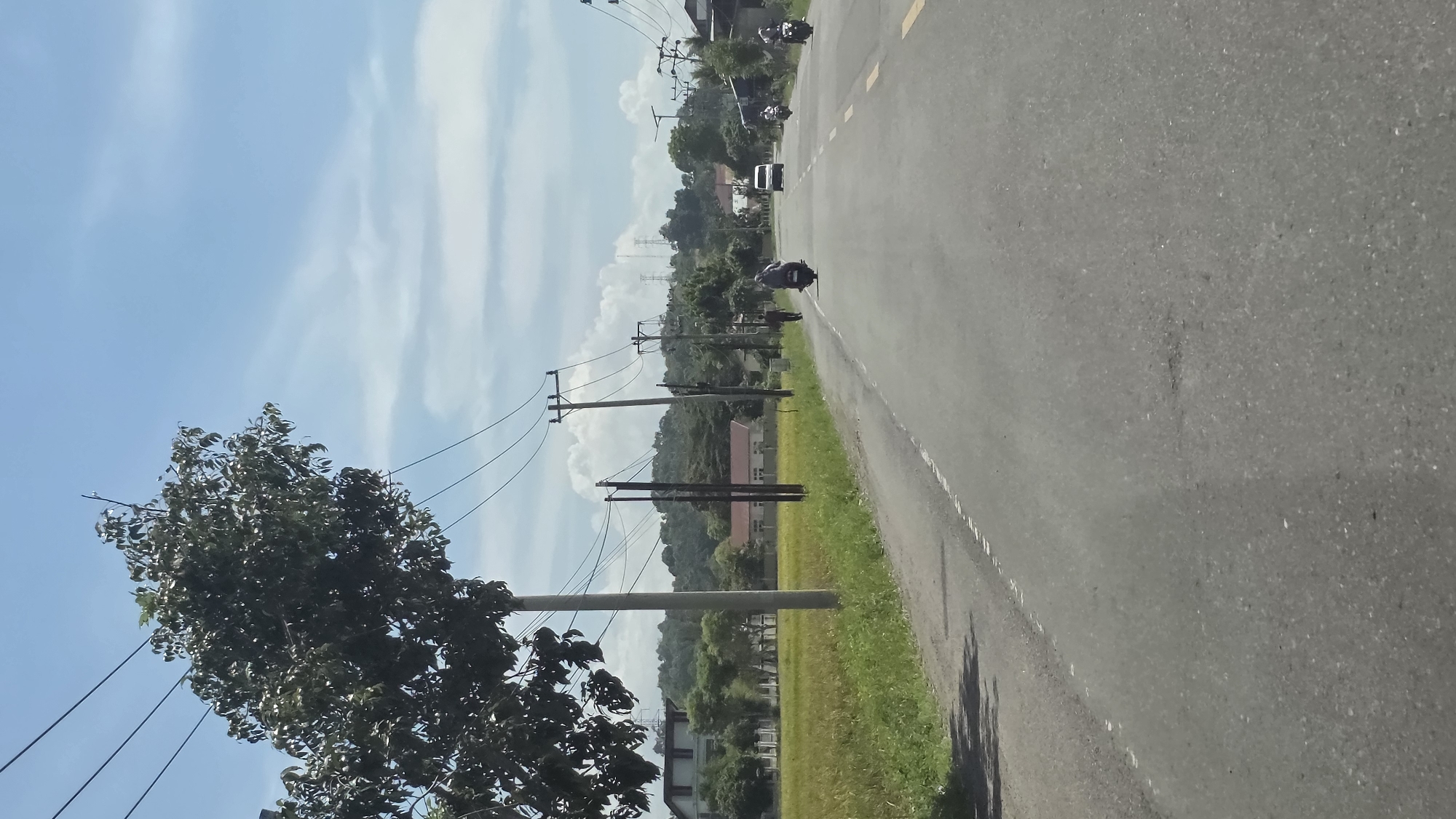 A roadside view in Manyang Cut, Indonesia, shows a gray asphalt road with a single white dividing line. Vegetation borders the road, with trees and grass. Power lines run along the road, supported by wooden poles. Motorbikes travel on the road. Buildings and hilly terrain are visible in the background, under a sky with scattered clouds.
