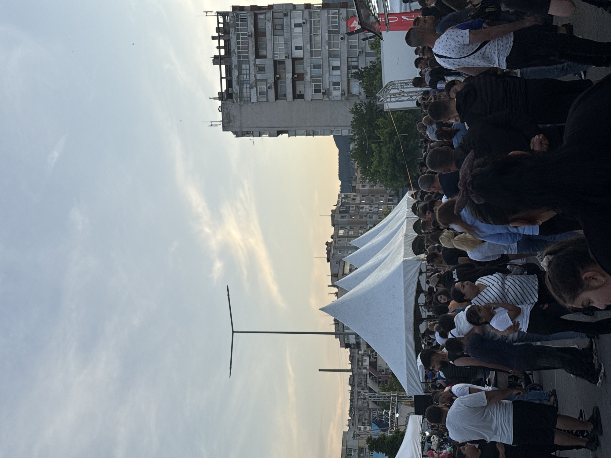 This image depicts a vibrant outdoor public gathering in Shumen, Bulgaria, during the late afternoon or early evening. The sky, a clear light blue with hints of warm light on the horizon and sparse, wispy clouds, indicates pleasant weather.
The scene is dominated by a large crowd of people, mostly adults dressed in casual attire such as t-shirts, shirts, and jeans. Many individuals have their backs to the camera or are seen in profile, appearing to be focused on a central point, likely a stage or speaker, which is partially visible on the right. Elements of a stage structure, including metal scaffolding and a red banner with a partial white letter "U" (possibly part of a word like "BULGARIA"), are discernible in the right background. A "NIKE" logo can also be seen on one person's shirt.
Several white, pointed event tents are prominent in the middle ground, suggesting an organized event such as a festival, concert, or public address. To the far left, a tall, slender pole with a T-shaped top stands against the open sky. In the background, typical Eastern European multi-story residential buildings with numerous windows and balconies line the cityscape. One taller building appears aged, with exposed rebar at its top, and distant hills or mountains complete the horizon line. The overall atmosphere seems attentive and calm, with the crowd engaged in the event.