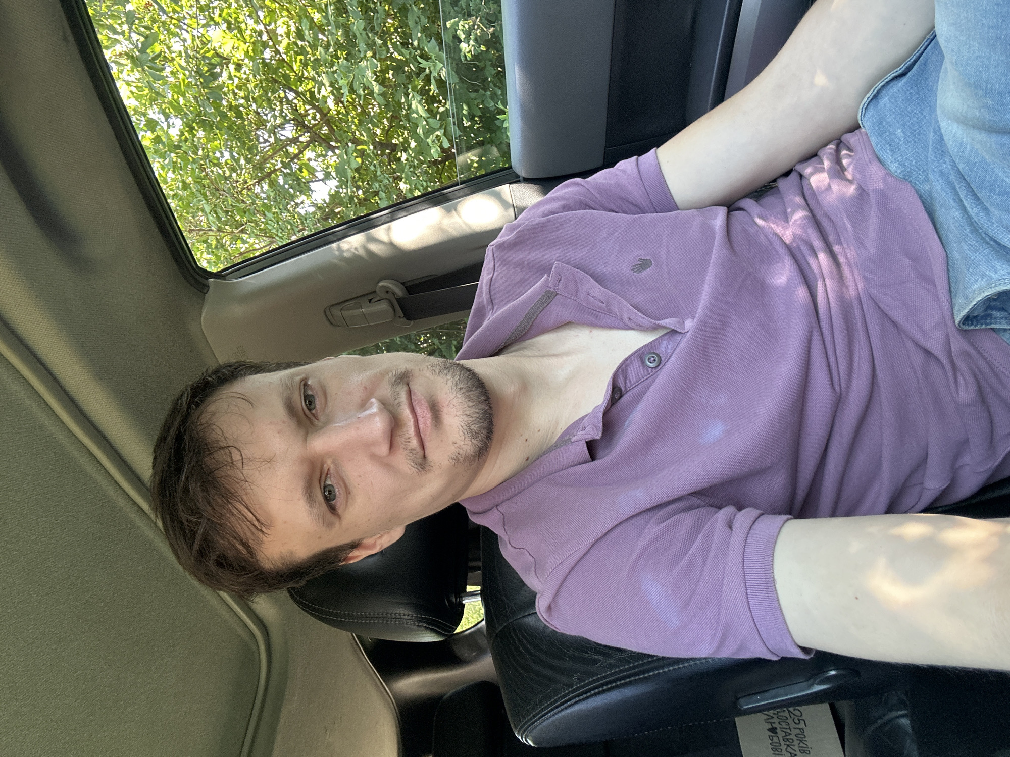 A man is sitting in the driver's seat of a car, facing the camera. He has light brown hair, blue eyes, and is wearing a purple polo shirt and blue jeans. The car's interior is visible, including the black leather seat and the headrest. Sunlight is streaming in through the open window, casting shadows on the man and the interior. Outside the window, lush green trees are visible. The scene appears to be in a rural or suburban area, given the presence of the trees and natural light. The location is Shatava, Ukraine.
