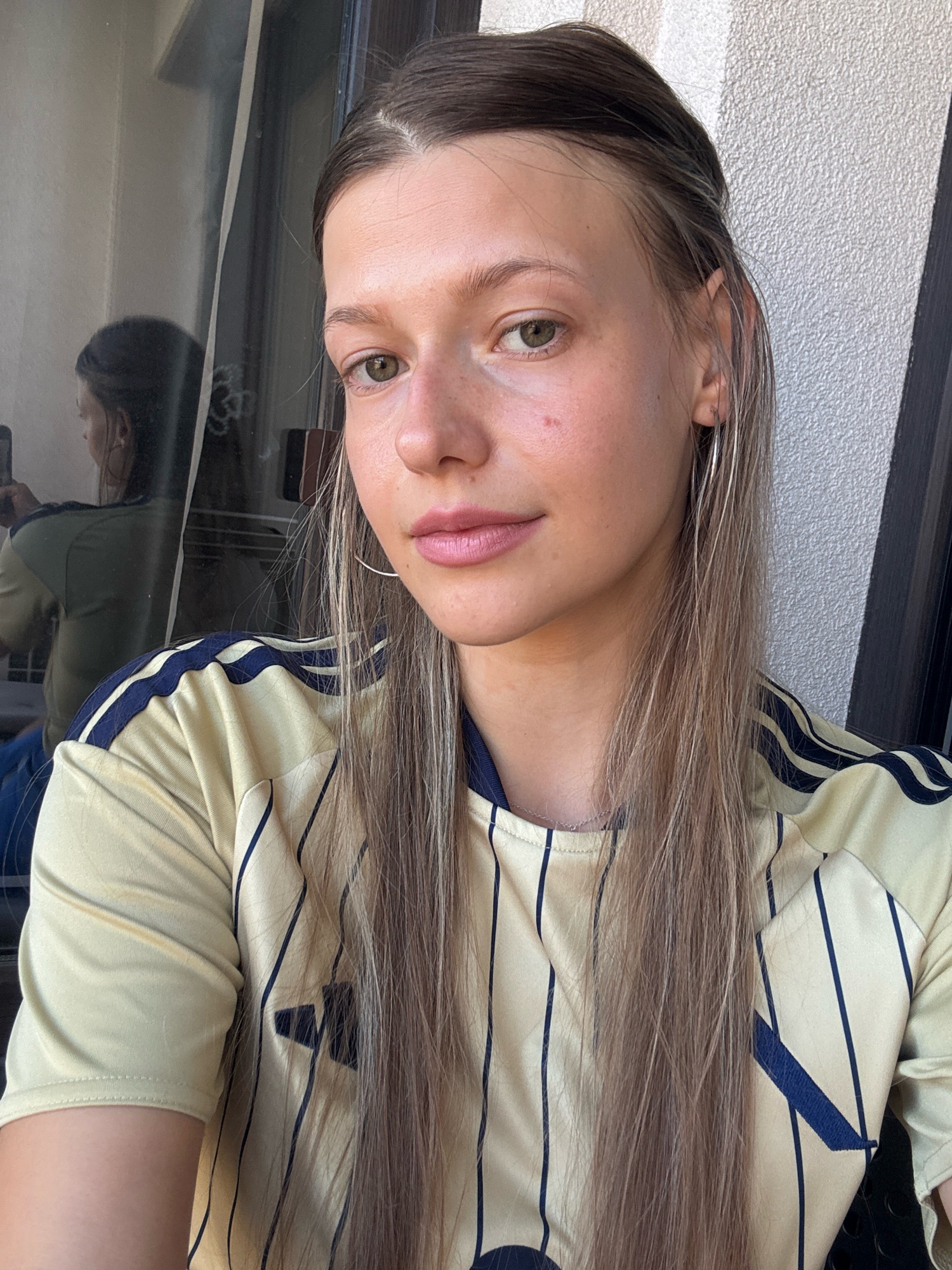 A young woman with long, blonde hair is pictured in a selfie. She is wearing a light yellow shirt with navy blue stripes and trim. The background shows a white textured wall and a dark framed window or door. A reflection of the woman in a mirror or glass surface is visible to her left. The image appears to have been taken outdoors, possibly on a balcony, given the presence of a railing and the natural light. There are no explicit indicators of her location within the city of Sofia, Bulgaria.