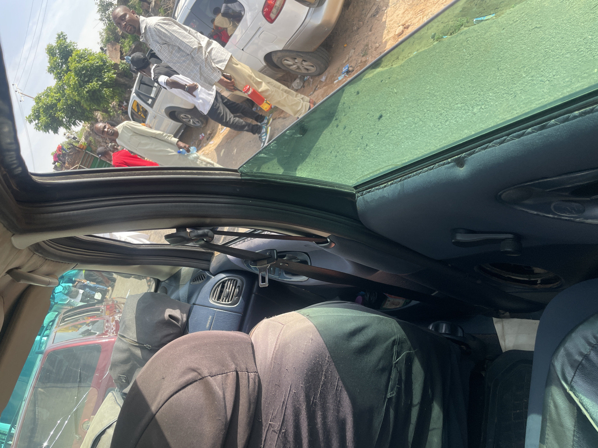 This image, taken from inside a vehicle in Kiri, Nigeria, shows a scene of activity on a dirt road. In the foreground, a person wearing a dark hooded jacket is seated in the passenger seat, with a dark duffel bag on their lap. The vehicle's interior features a blue dashboard with an air vent and a seatbelt. Through the car's side window, several people are visible standing and interacting outdoors. A man in a light-colored tunic holds a water bottle, while another man in a plaid shirt stands nearby. Several vehicles, including a white van and a red car, are parked on the roadside, suggesting a gathering or a temporary stop. The environment appears to be somewhat rural, with a dusty road and vegetation in the background. The overall impression is of a moment captured during a journey or a stop in a Nigerian community.