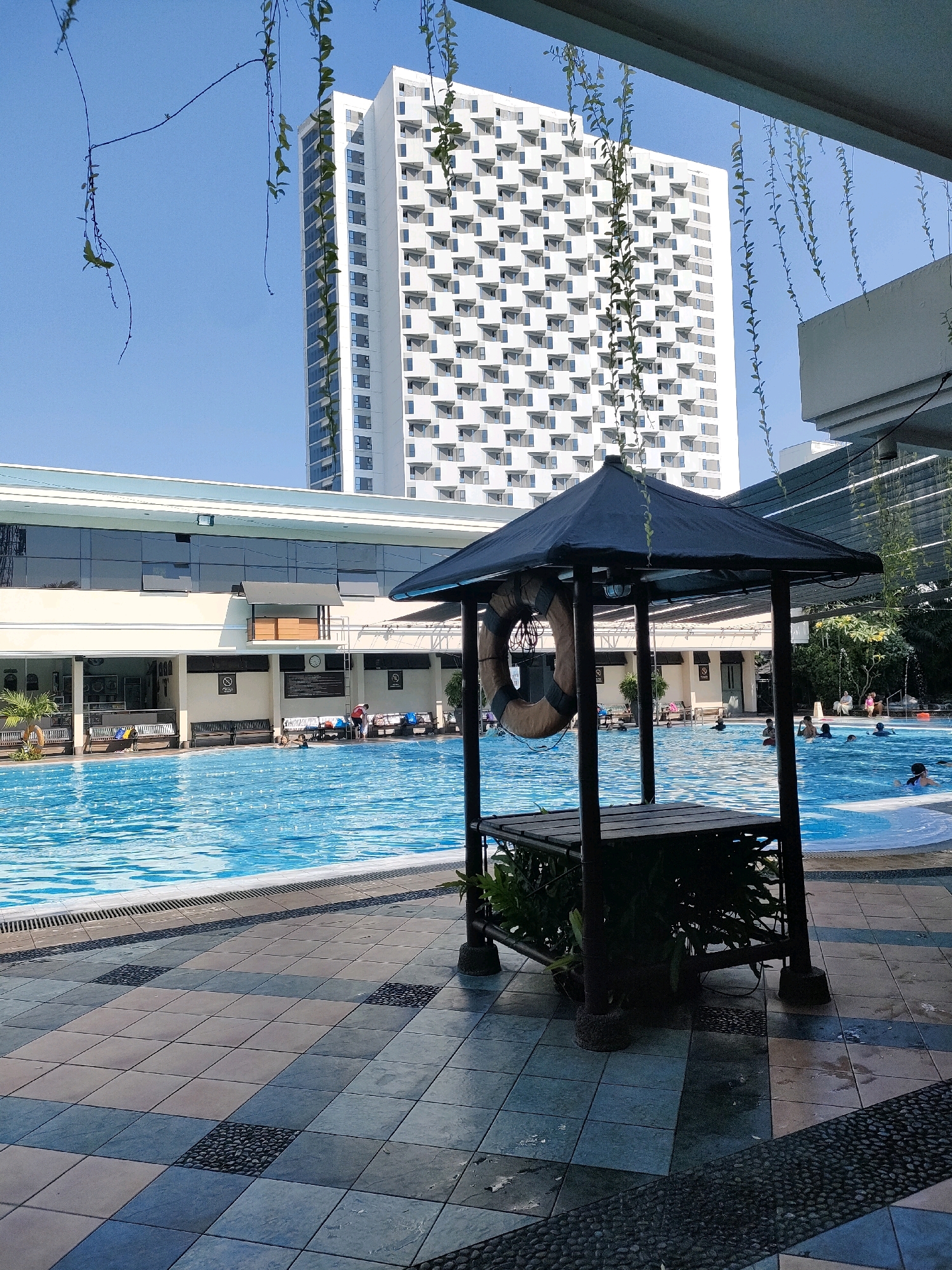 This image depicts a sunny outdoor swimming pool area, likely at a hotel or resort, in Surabaya, Indonesia. In the foreground, a dark-colored gazebo with a wooden table and hanging plants stands by the pool's tiled edge. A life preserver hangs on one of the gazebo's posts. The pool itself is a vibrant blue, with ripples on the surface and several people swimming and relaxing.
Behind the pool, a long, low-rise building with a white facade and numerous windows runs along one side. Further in the background, a very tall, modern white high-rise building with a repeating pattern of square windows dominates the skyline. The sky is clear and blue. The pool area is paved with multi-colored square tiles, interspersed with darker stone sections. Green vines hang down from above, partially obscuring the view of the buildings and sky. The overall impression is one of a pleasant, recreational outdoor space within an urban environment.