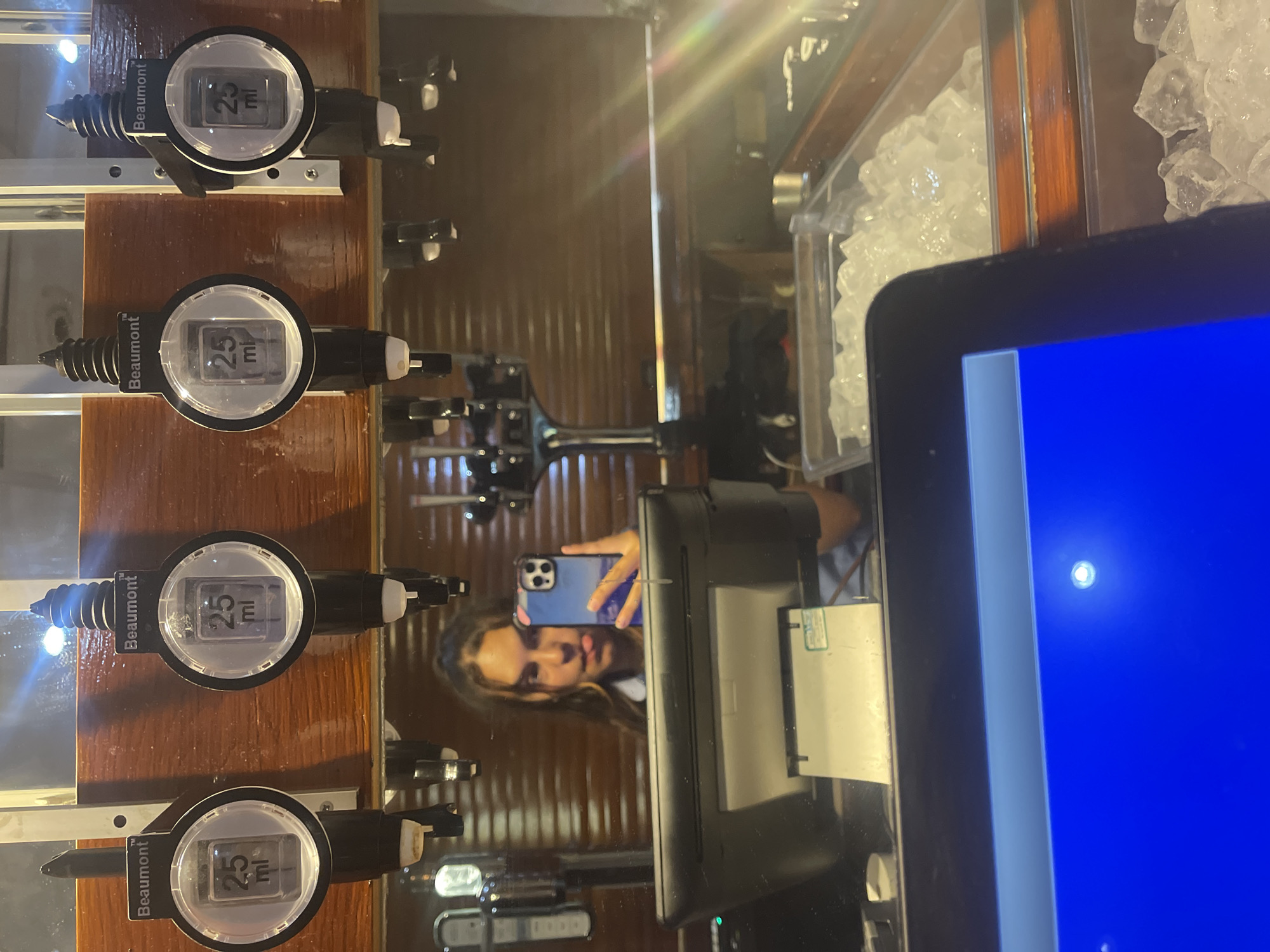 The image captures a mirrored reflection of a young woman at a bar in Maidstone, United Kingdom. She is taking a selfie with her phone, sticking her tongue out playfully. Behind her, a row of four Beaumont brand liquor dispensers is visible, each set to 25 ml. The bar features a wooden paneling background, and in the foreground, a touch-screen POS system and a container filled with ice cubes are present. The overall scene suggests a casual, perhaps evening, setting in a hospitality establishment.
