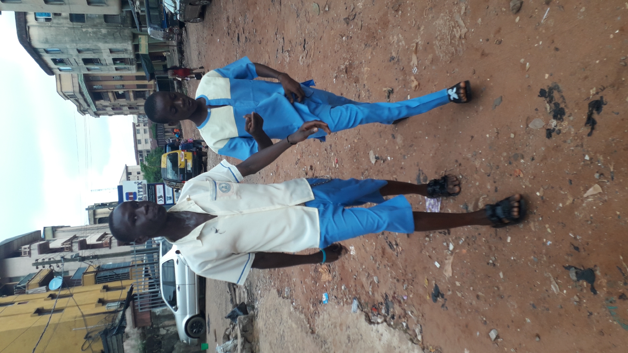 Two young African men stand outdoors on a dirt path in Onitsha, Nigeria. Both wear sandals. One wears a long-sleeved, two-tone blue and cream uniform, and the other wears a short-sleeved version. Urban buildings line the background, alongside passing cars, motorcycles and a taxi. There is trash on the ground.
