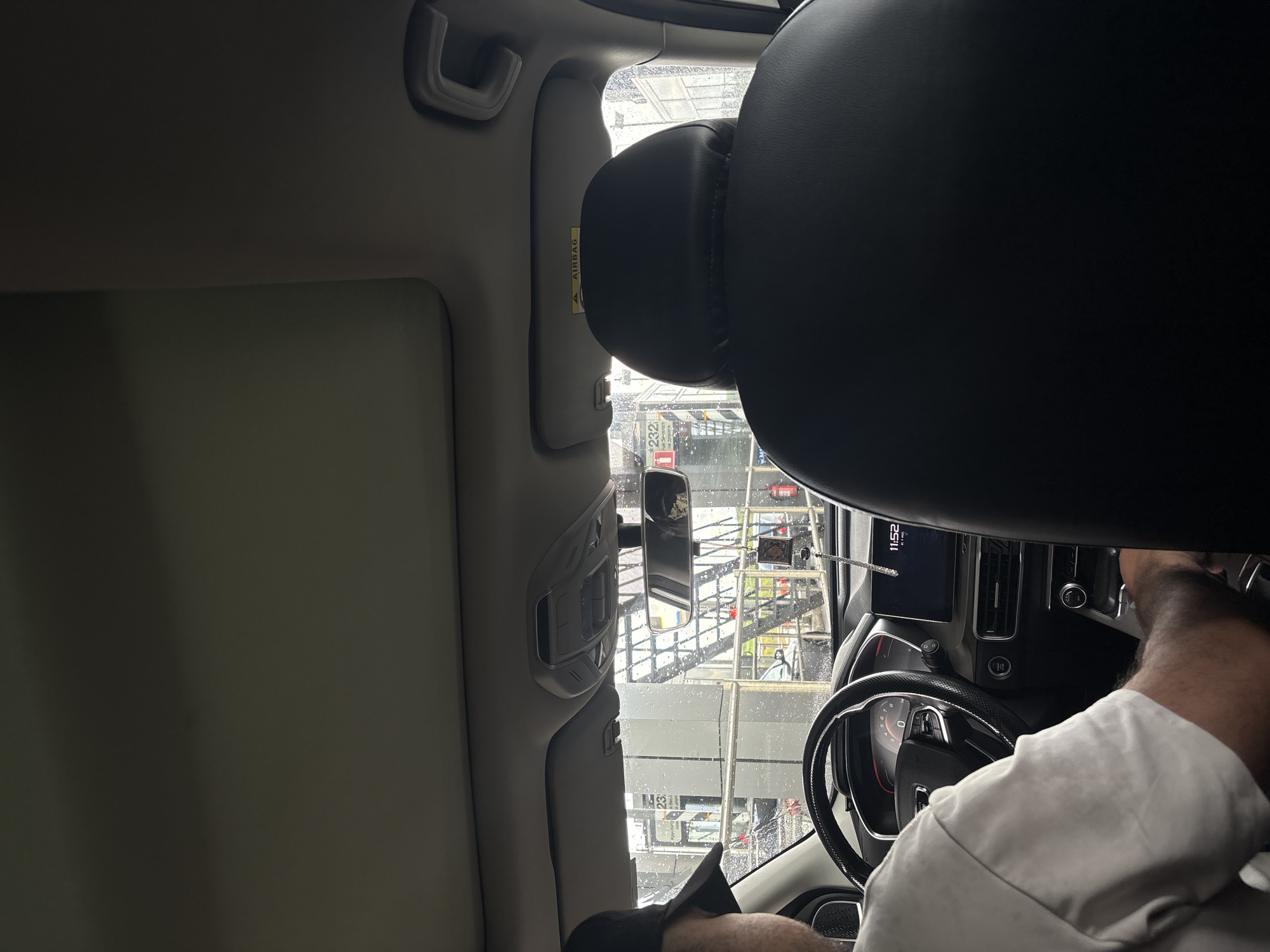 The image shows the interior of a car, viewed from the back seat, looking towards the front. A person wearing a white t-shirt and a black baseball cap is seated in the driver's seat, with their left arm resting on the steering wheel. The car's dashboard and infotainment screen are visible. The infotainment screen displays the time as 11:52. Outside the car, through the windshield, a fuel station is visible. Specifically, it appears to be a Lukoil fuel station in Khimki, Russia, as indicated by the presence of the Lukoil logo and Russian text on the fuel pumps and signage. The weather is overcast and it appears to be raining, with visible raindrops on the windshield.