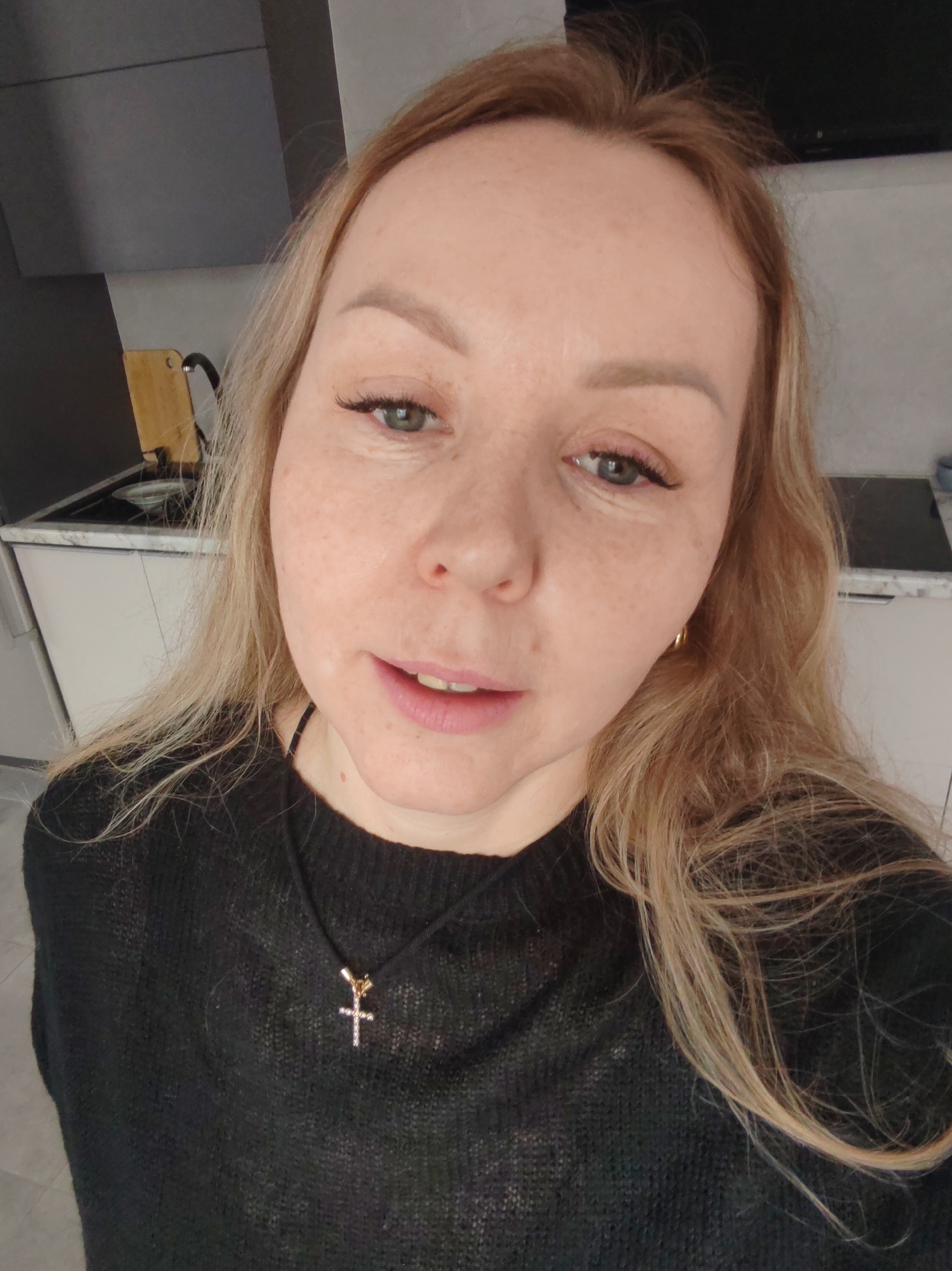 A selfie of a woman with light blonde hair and green eyes wearing a black sweater and a cross necklace. The background shows a kitchen with white cabinets and a grey countertop. Based on the prompt, it is located in Chaikovsky, Russia.