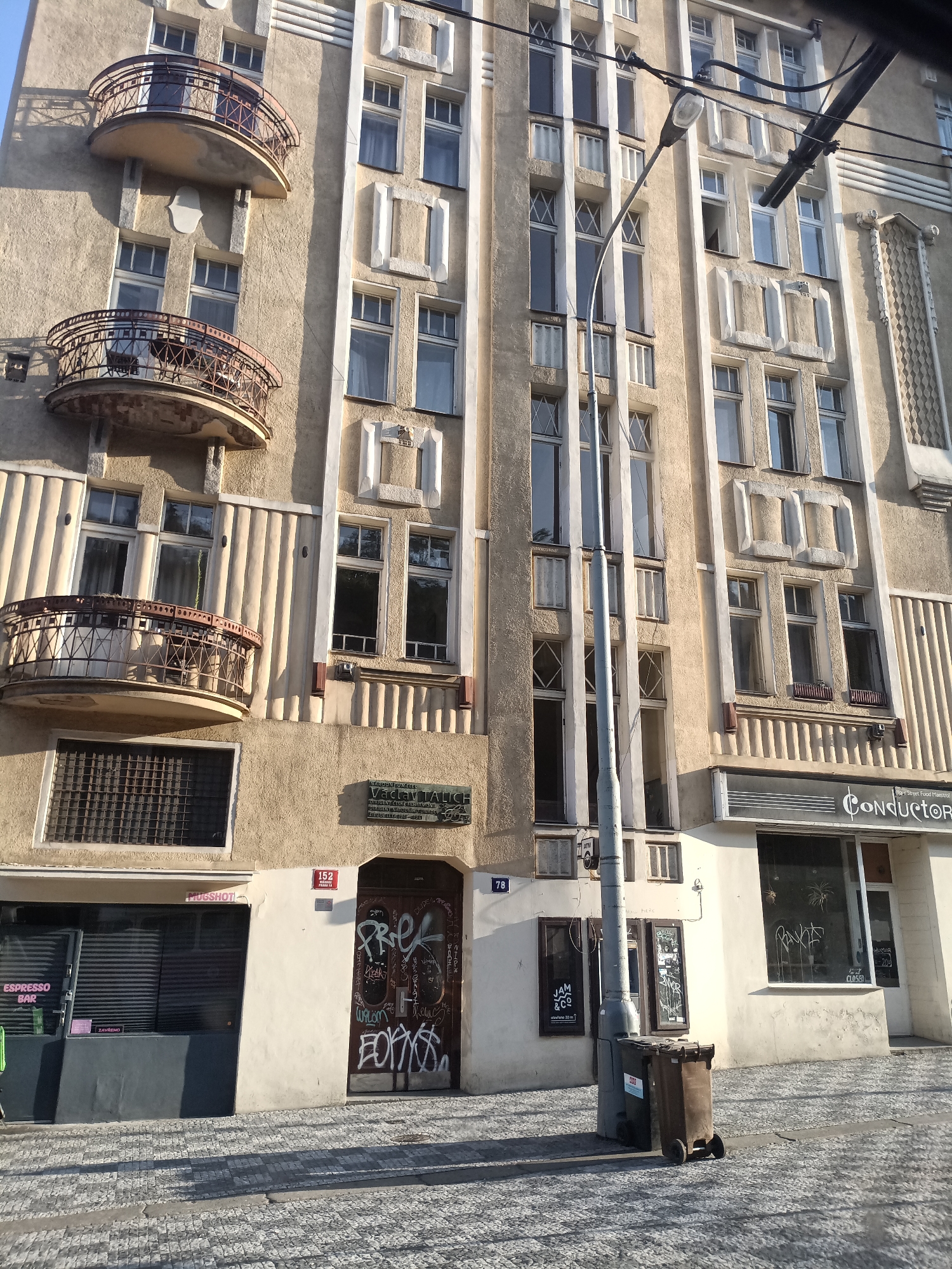 The image shows the exterior of a building in Prague, Czechia. The building is tall with multiple floors and features a pale yellow facade with decorative white trim. There are several balconies with ornate metal railings. The windows are arranged in a grid pattern, with some windows featuring a criss-cross design.
At street level, there are several businesses. On the left, there is a store with a dark gray facade and the text "ESPRESSO BAR" and "THUGSHOT" visible. The windows are covered with blinds. Next to it, a brown door is covered in graffiti. Above this door, a plaque is affixed to the wall, indicating "NA PŘEDNÁŠKU VACLAV TALICH" (For the lecture Václav Talich). To the right of the graffiti-covered door, there is a business with a large display window that has the text "CONDUCTOR" and "Street Food Maestro." Some of the windows are covered with graffiti. A lamppost stands in front of the building, and a trash bin is located next to it. The ground is paved with cobblestones. The sky is visible at the top of the image and appears to be clear.
