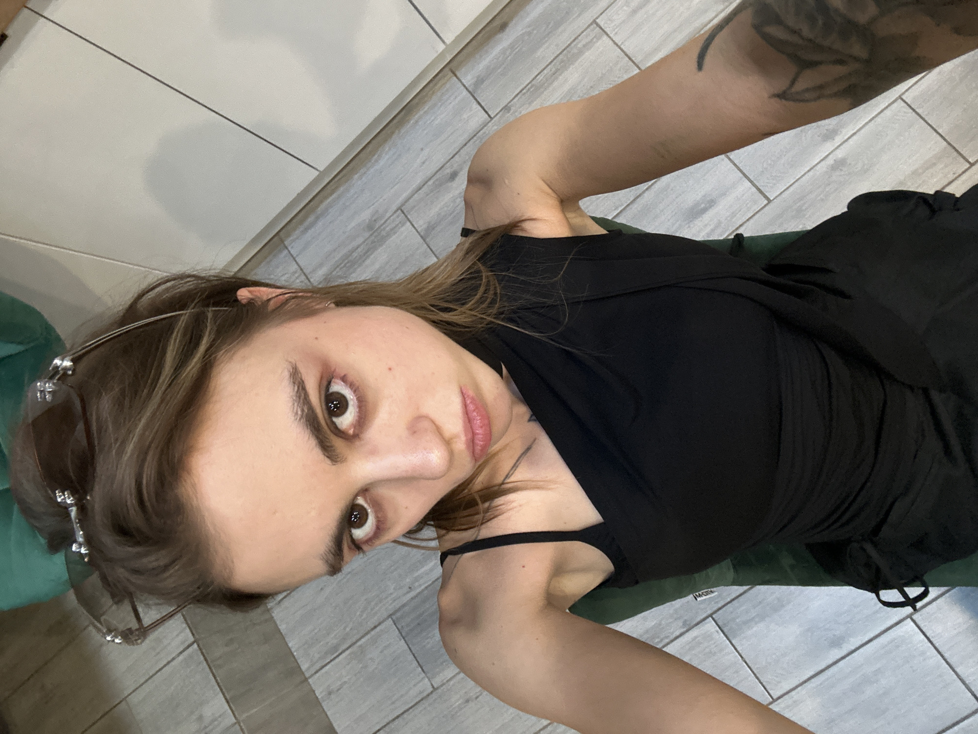 A young woman with brown hair and brown eyes is looking directly at the camera with a pout. She is wearing a black dress with an asymmetrical neckline. She has makeup on her eyes, including dark eyeshadow. Her eyebrows are dark and full. She has a tattoo on her left arm. She is sitting down, and a green surface is visible beneath her. The background appears to be a floor with light-colored tiles. There are also some white cabinets or walls in the background. A pair of sunglasses is resting on top of her head. The overall impression is a candid selfie taken indoors. There is no information about the location in Moscow, Russia.