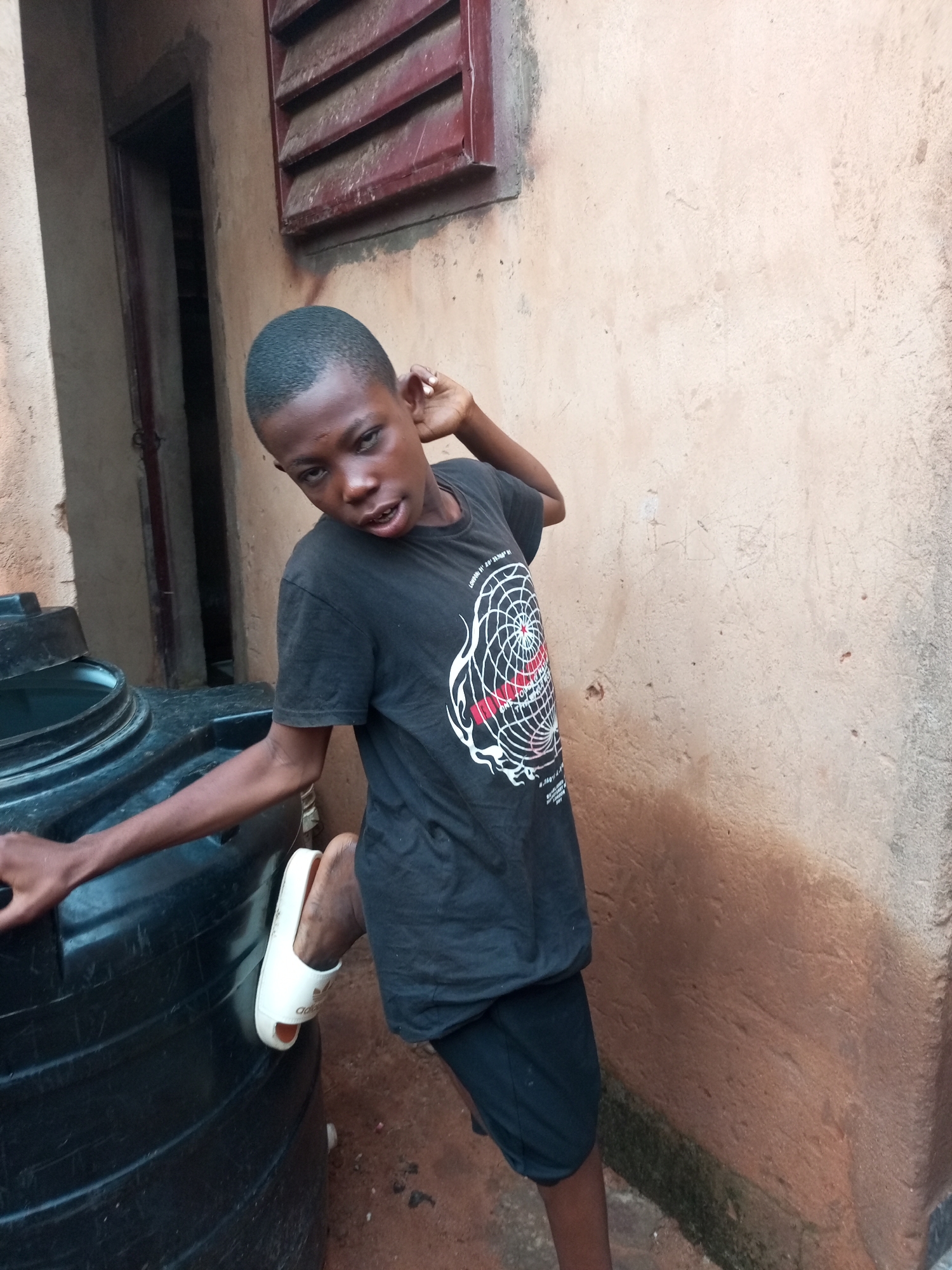 A young Black boy, wearing a black t-shirt and black shorts, stands next to a large black plastic water tank outdoors. He is leaning his left elbow on the tank and his right hand is behind his head. His left foot is resting on the side of the tank, and he is wearing a white Adidas slipper on that foot. The boy is looking towards the camera with an open mouth. The background consists of a textured, light brown wall and a dark doorway with a partially visible window with red shutters above it. The scene appears to be in a residential area in Tempure, Nigeria.