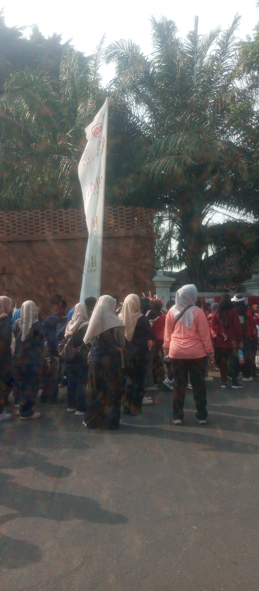 In Kediri City, Indonesia, a group of people, mostly women wearing hijabs, are gathered outdoors. The scene appears to be a public space, possibly near a building with a brick facade and a tall white flag on a pole. Palm trees are visible in the background, suggesting a tropical or subtropical climate. The people are standing and facing away from the camera, their attention seemingly directed towards something out of frame or the flag. One woman in the foreground is wearing a bright orange long-sleeved top and dark pants, while others are dressed in darker, more muted colors. The ground is paved, and shadows are cast by the people and possibly nearby structures, indicating it is daytime. The overall atmosphere seems to be one of a gathering or event.