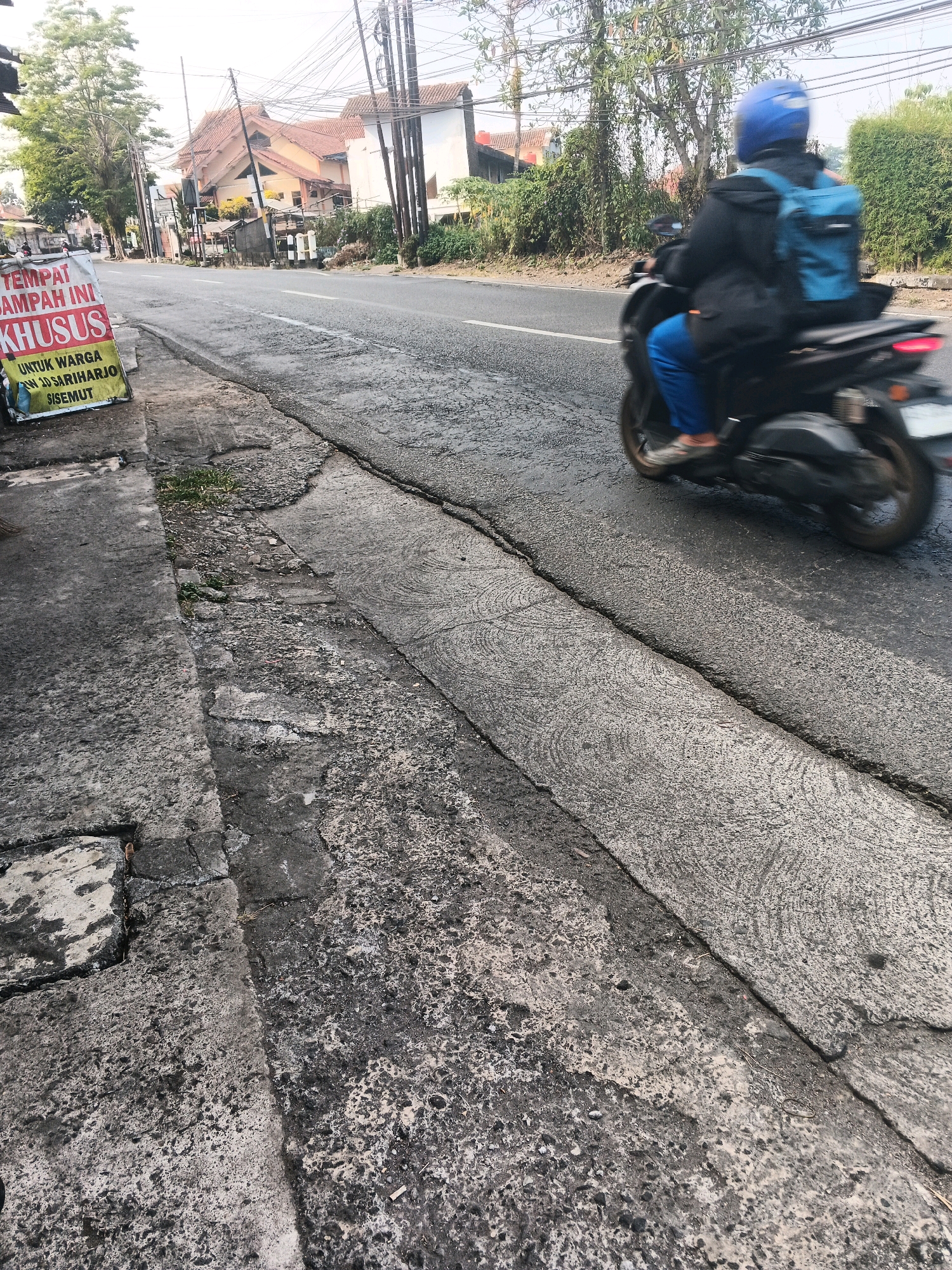 In Ungaran, Indonesia, an individual on a motorbike, wearing a blue helmet and a blue backpack, travels down a street. The road is paved and appears to be in moderate condition, with visible cracks and textured patterns on the surface. To the left of the road, a concrete sidewalk or gutter area is present, showing signs of wear and tear. A sign on the left side of the image reads "TEMPAT SAMPAH INI KHUSUS UNTUK WARGA RW 10 SARIHARJO SISEMUT," indicating a designated trash disposal area specifically for residents of RW 10 Sariharjo Sisemut. In the background, buildings with tiled roofs and trees line the street, suggesting a semi-urban or residential area. Overhead, numerous electrical wires are visible, a common sight in Indonesian cities. The overall scene depicts a typical street in Ungaran during daylight hours.