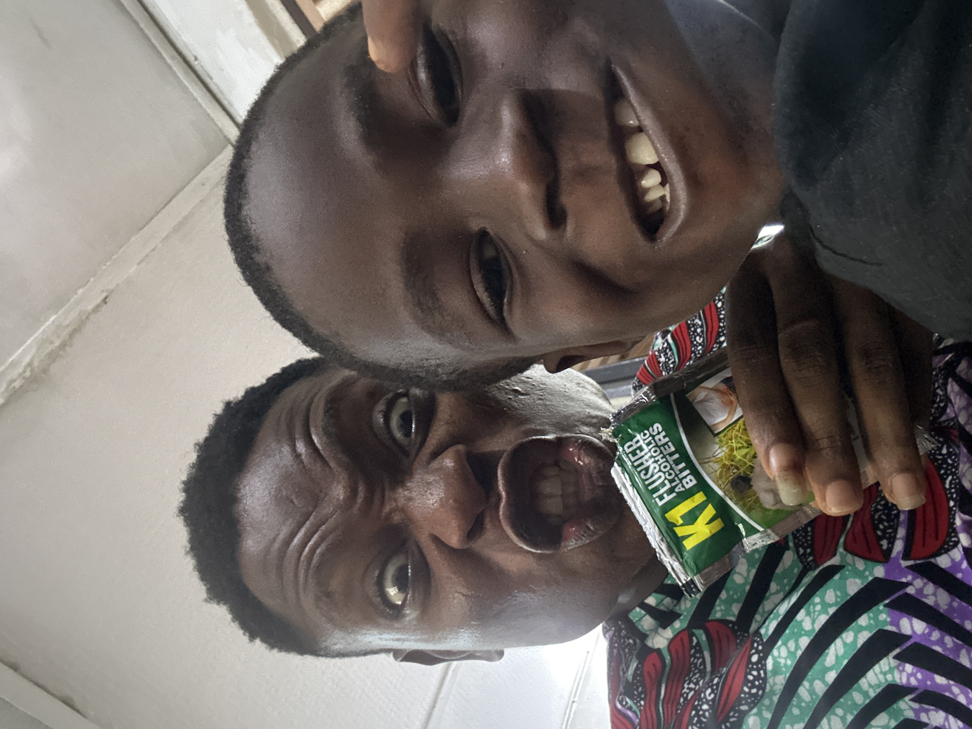Two dark-skinned individuals are captured in a close-up, angled selfie. The person at the top is grinning, while the one below is making a distorted facial expression, holding a packet labeled "K1 Flusher Alcoholic Bitters." The person holding the packet is wearing a colorful shirt with patterns. The background suggests an indoor setting, possibly a room with a white wall. The city and location are unknown.