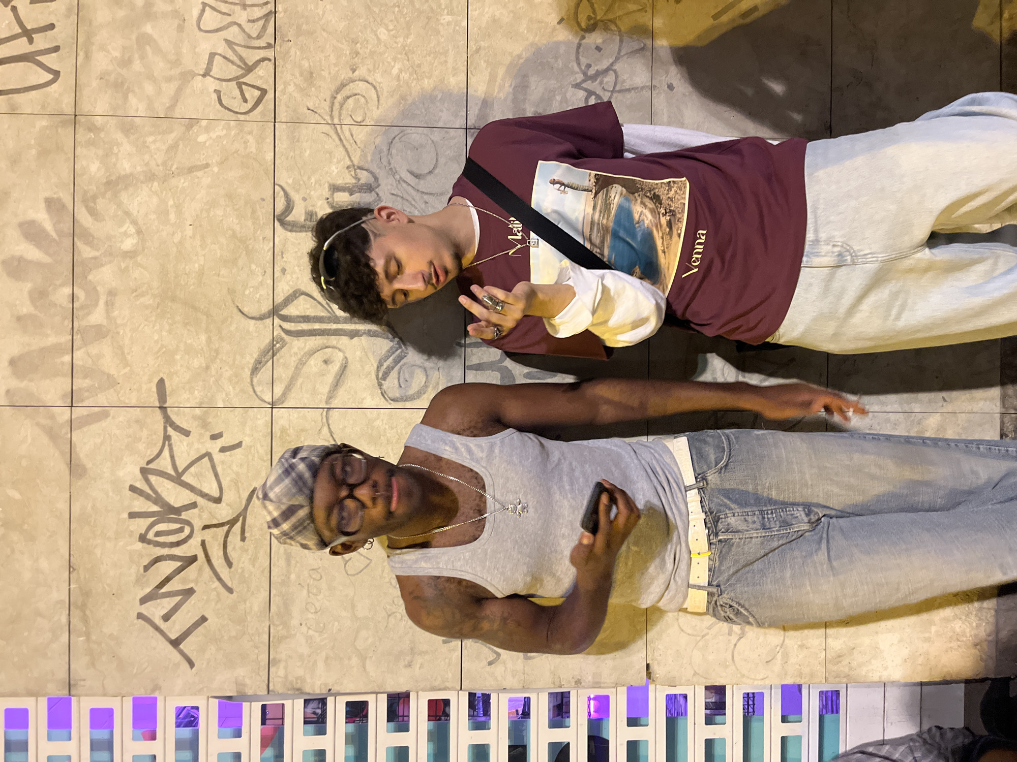 Two young men are lying on a tile-covered surface outdoors. The tiles have graffiti on them. One man is wearing a burgundy shirt with a picture and the word "Vanna" on it, light-colored jeans, and a shoulder bag. He is lying on his back with his eyes closed and has multiple rings on his fingers. The other man is wearing a gray tank top, light-colored jeans with a white belt, glasses, and a patterned cap. He is also lying on his back and is holding a small object in his hand. There is a row of decorative white and blue columns at the bottom of the frame. The location appears to be an urban area, likely Barcelona, Spain, given the tile work and the styles of dress.