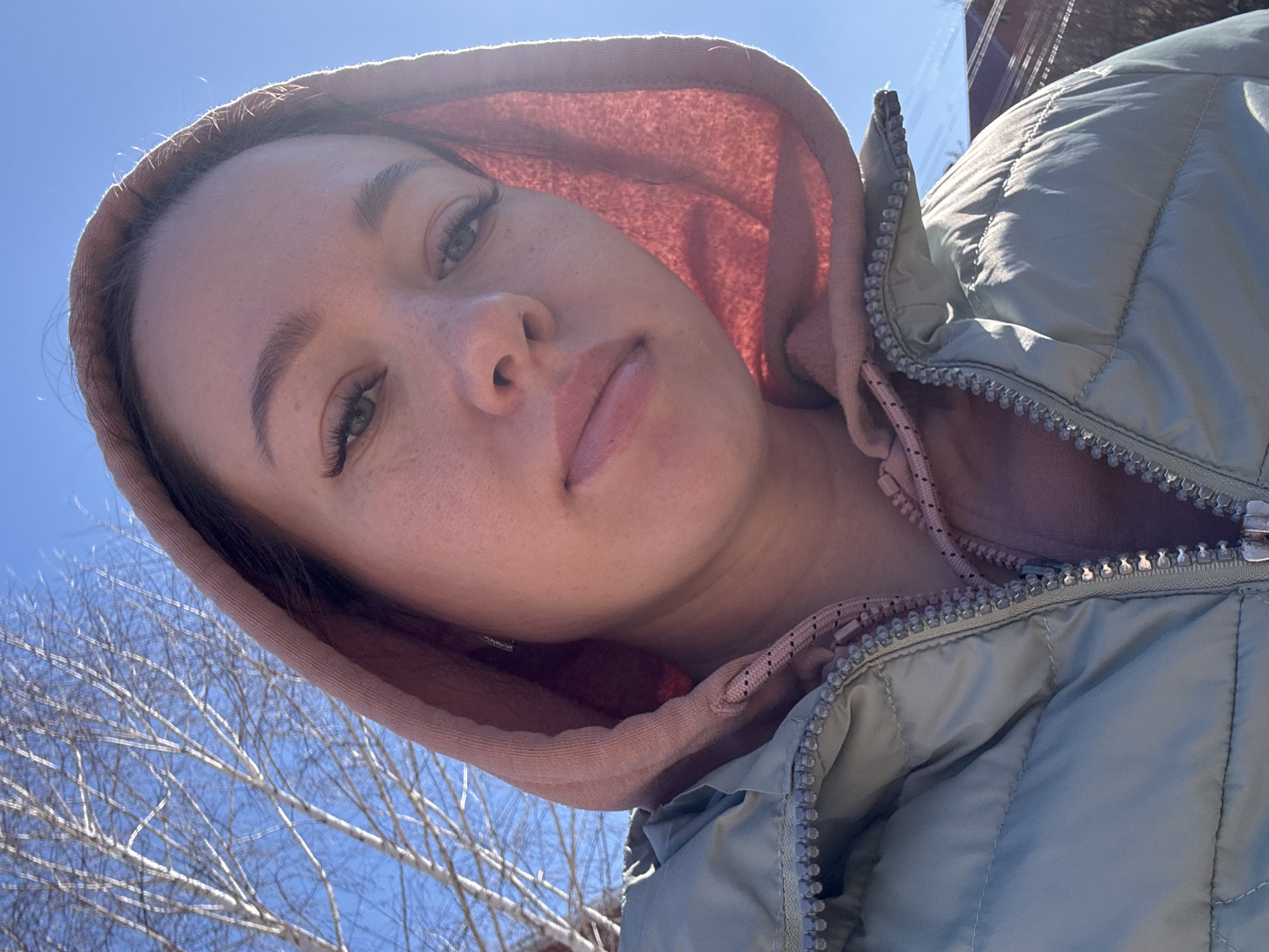 A woman with light skin, freckles, and light-colored eyes looks down at the camera. She is wearing a puffy, light green coat with a pink hoodie underneath. The hoodie is up. The sky is blue and there are bare tree branches in the background. The photo appears to have been taken outdoors during the day. Given the location, it is implied that the image was taken in or near Ферма, Russia.