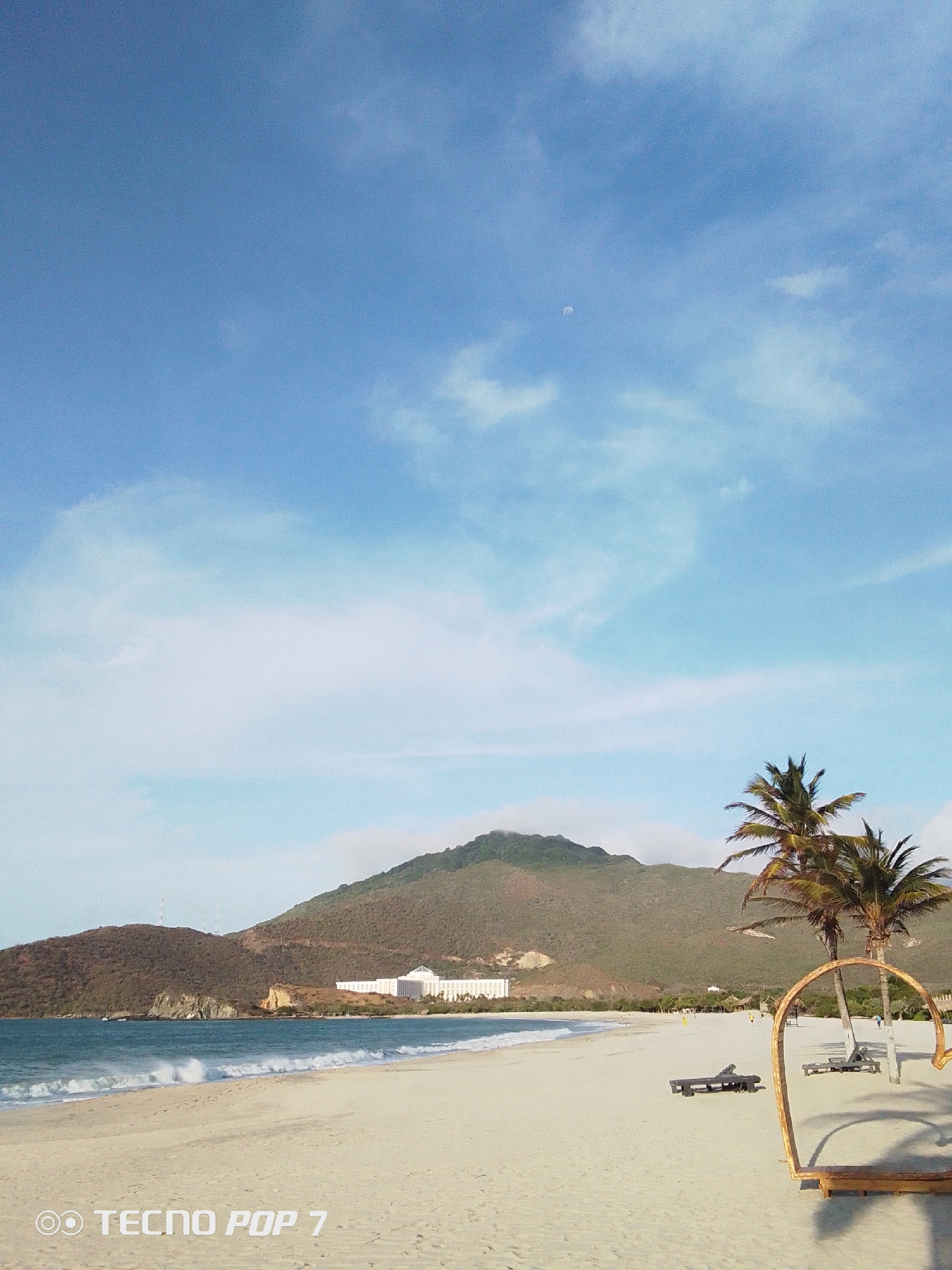 The image shows a coastal beach scene in Pedro González, Venezuela. The foreground consists of a sandy beach with scattered palm trees and wooden structures. Two deckchairs are visible on the sand. Behind the beach, a body of water meets the shore with small waves. In the background, hills and a mountain dominate the horizon. There is a large, light-colored building situated along the coastline. The sky is mostly blue with scattered white clouds and a visible moon. The image also includes a watermark at the bottom left corner, indicating that it was taken with a "TECNO POP 7" camera.
