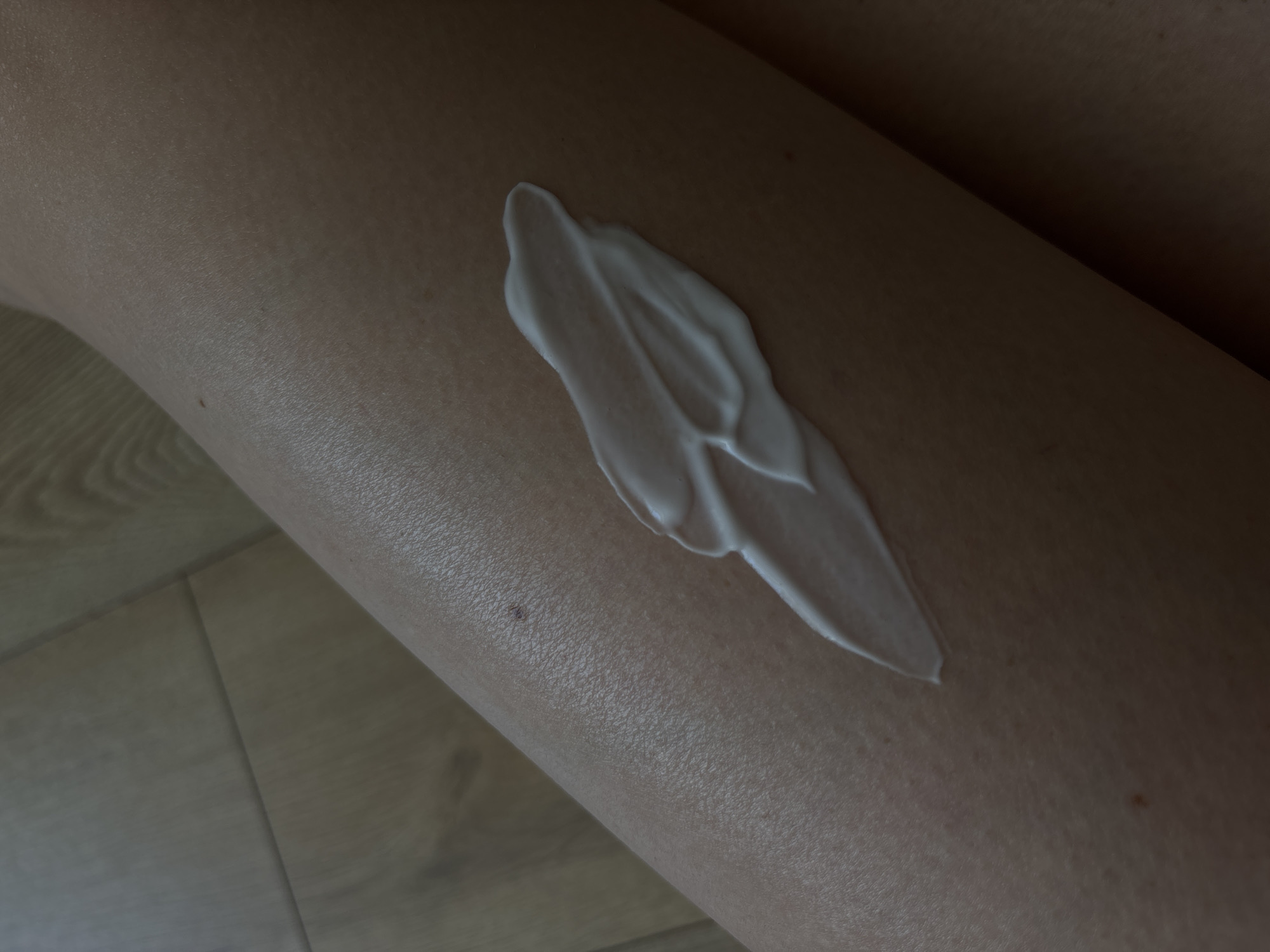 This is a close-up, overhead shot of a person's leg with a streak of white lotion applied to it. The skin is fair and appears dry, with visible pores and a few small moles or blemishes. The lotion is thick and creamy, applied in a sweeping motion, creating a smooth, opaque contrast against the skin. The background is out of focus, showing a wooden floor with a light-colored grain pattern. The lighting appears to be artificial, casting a warm, soft glow on the scene. There is no visible text in the image. The overall impression is one of personal care or a beauty routine. The focus is on the texture of the skin and the application of the lotion, highlighting a moment of self-care or skincare.