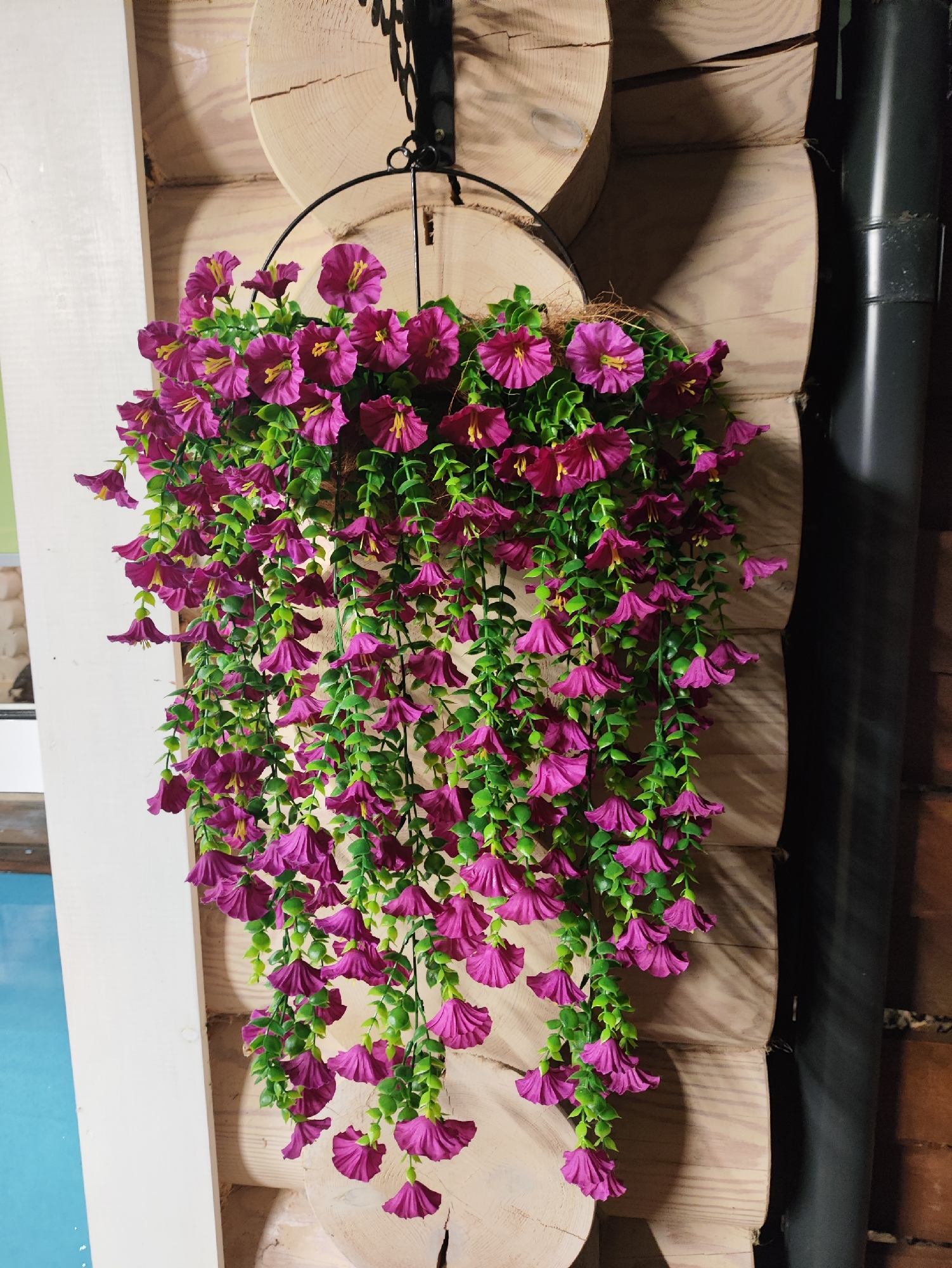A hanging basket filled with artificial purple flowers with yellow centers and green leaves is suspended against a backdrop of light-colored, rough-hewn logs. The logs appear to be part of a rustic structure, possibly a wall or a decorative element. To the left of the logs, a portion of a white structure is visible, and to the right, a dark, cylindrical object, possibly a pipe or a downspout, runs vertically. The overall scene suggests an outdoor or rustic indoor setting. The location is specified as Staroye Martyánovo, Russia, but there are no visible elements in the image that specifically confirm this location. There are no people or animals present in the image.