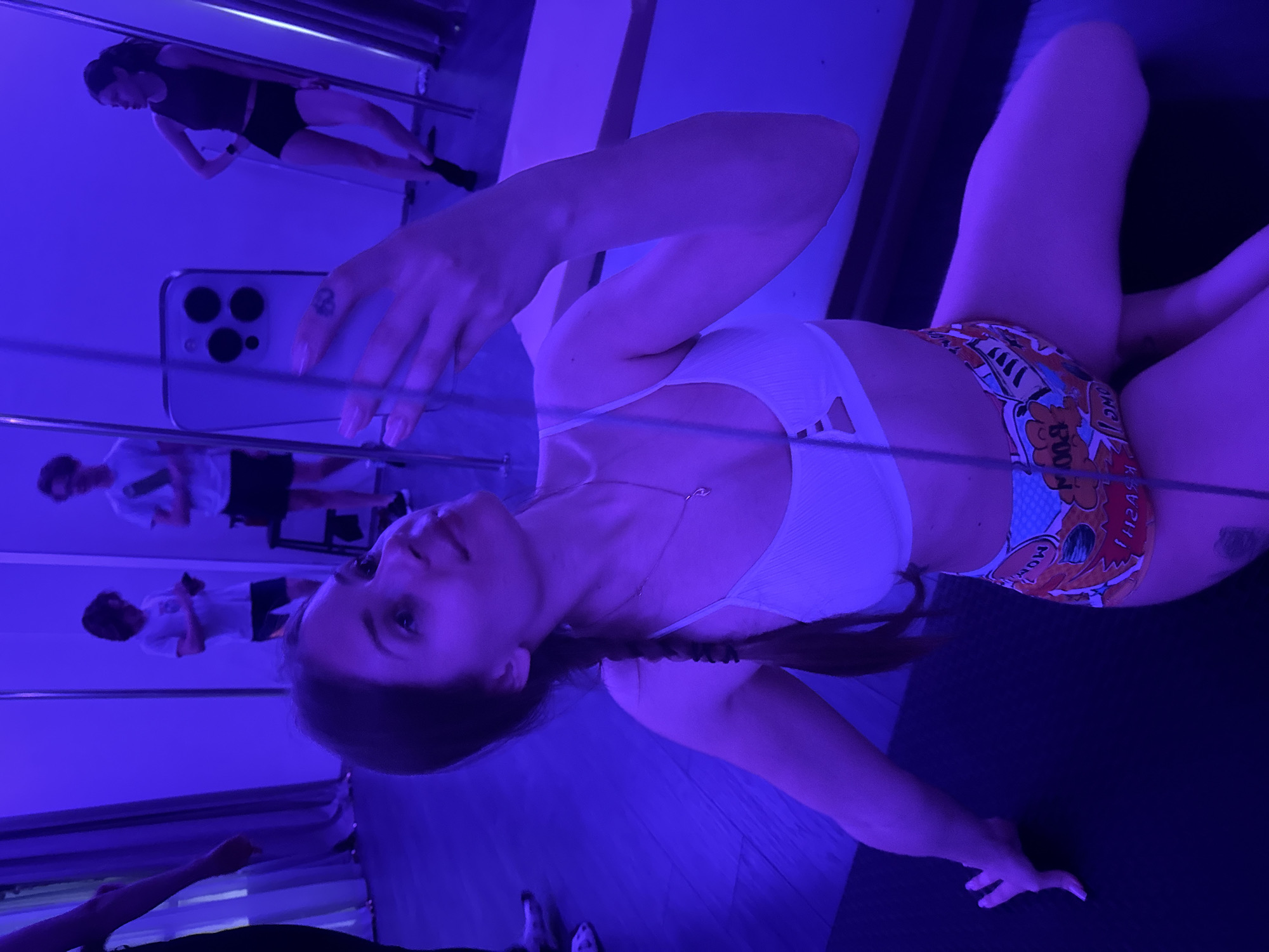 A woman is taking a selfie in front of a mirror. The lighting is a strong blue hue. She is wearing a white sports bra and patterned shorts. In the mirror's reflection, there are other people, one in black athletic wear and others in white shirts and dark shorts. The specific location in Batumi, Georgia cannot be determined from the visual information provided.