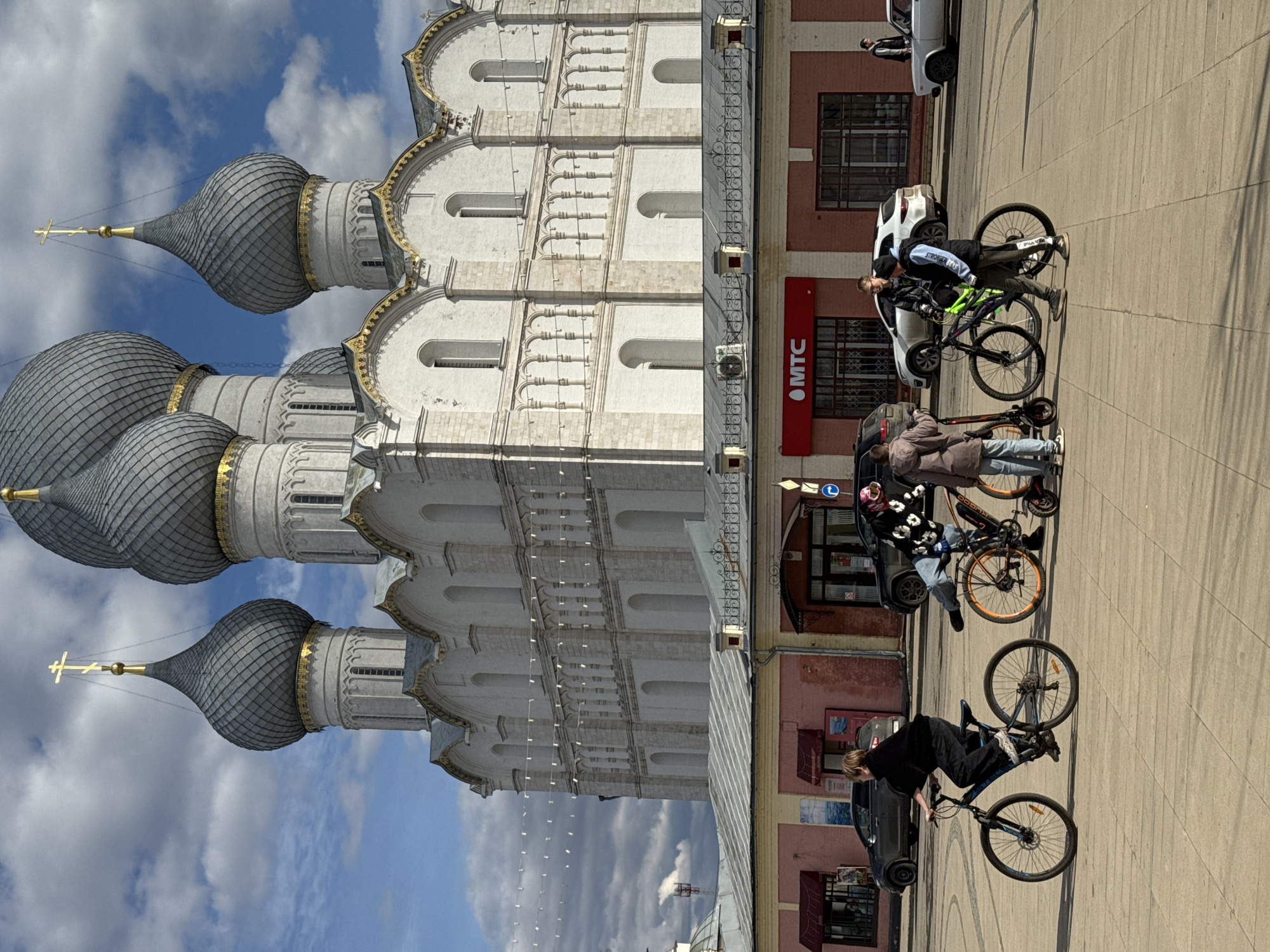 The image depicts a street scene in Семибратово, Russia, featuring the Rostov Kremlin in the background. The Kremlin is a large, white stone building with several onion-shaped domes topped with crosses. In the foreground, there are four people riding bicycles on the sidewalk. Several cars are parked along the street adjacent to the sidewalk. A storefront with the red "MTC" logo is visible on a building near the cyclists. The sky is blue with scattered clouds.