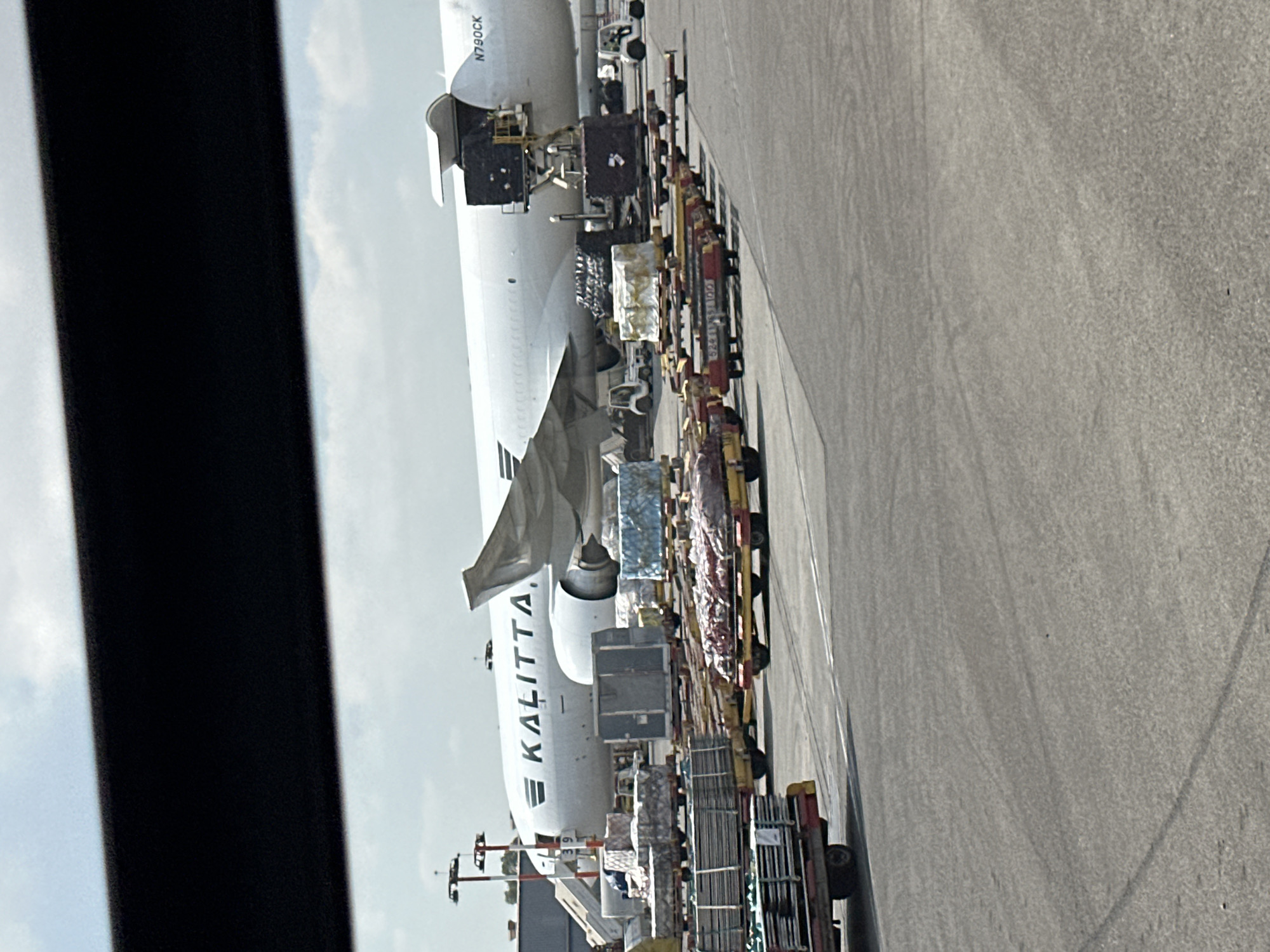 The image shows an aerial cargo plane with the Kalitta Air logo on its side. The plane is parked on an tarmac, with its cargo door open and containers being loaded or unloaded. There are several cargo trolleys with various goods on them, as well as small towing vehicles. The scene appears to be an airport or an air cargo facility. The specific location in the city of Zeitan, Israel, is not identifiable from the image itself. The weather appears to be clear with some clouds in the sky.