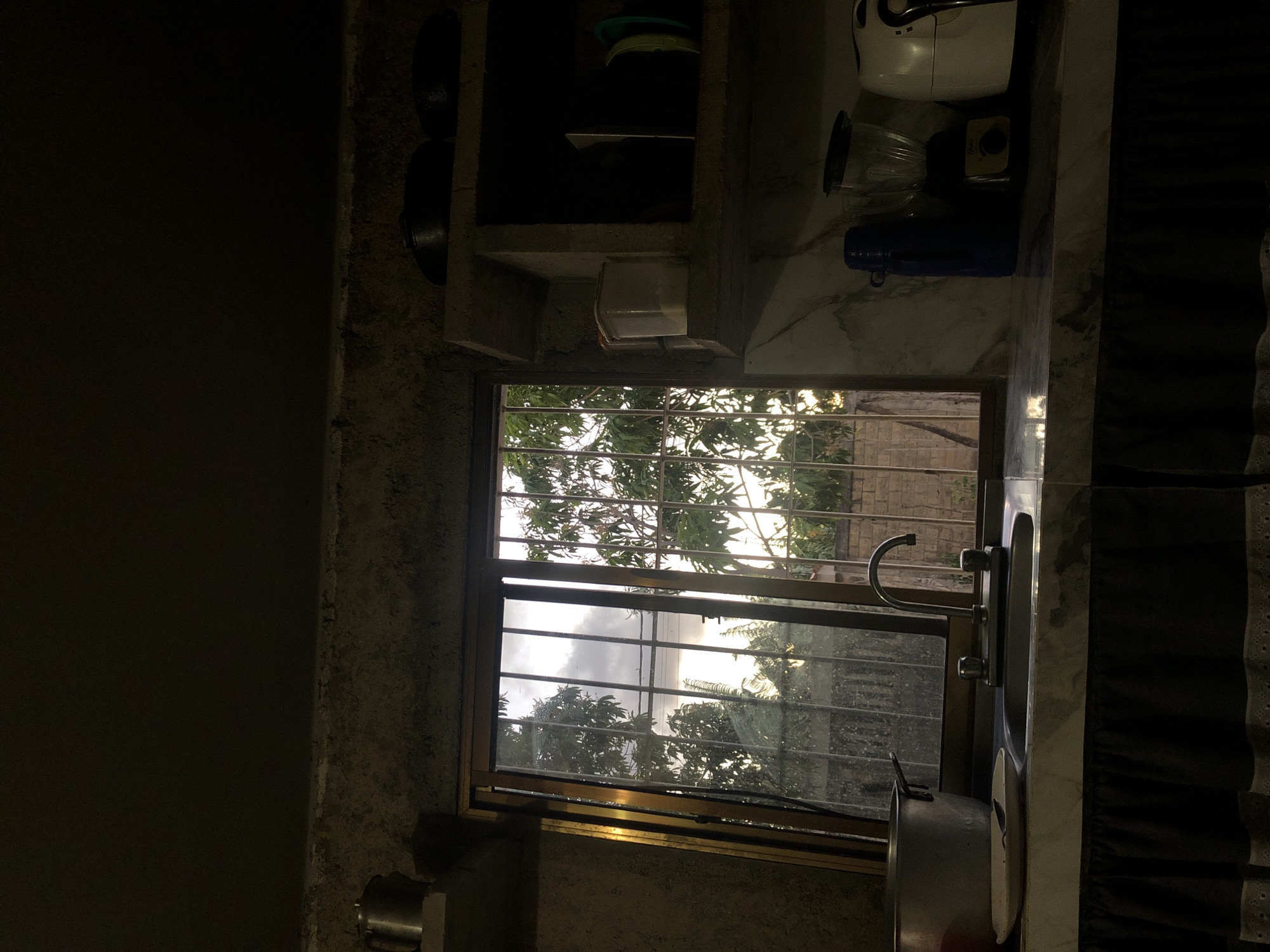 The image depicts a dimly lit kitchen interior, likely in Altagracia, Venezuela, viewed towards a window. The room features rough, textured concrete or stucco walls and ceiling, with minimal direct light. A double-basin sink with a gooseneck faucet is centered in front of a rectangular window, which is fitted with vertical security bars and a metal frame. To the left of the sink, a large silver-colored pot and a white plate with a dark utensil rest on the marbled countertop. To the right, a blue thermos, a blender, and a white kitchen appliance (possibly an air fryer) are positioned. Above the right side of the counter, built-in concrete shelves hold various items, including a clear plastic container with white contents and several dark pots or pans. A similar shelf on the left holds a metal pitcher. Below the countertop, a dark fabric skirt with a white lace or embroidered trim is visible. Through the window, lush green foliage from trees, possibly mango trees, and a concrete block wall are visible under an overcast sky, suggesting either dawn or dusk. The exterior light brightly illuminates the trees, contrasting with the dark interior. No individuals are present.