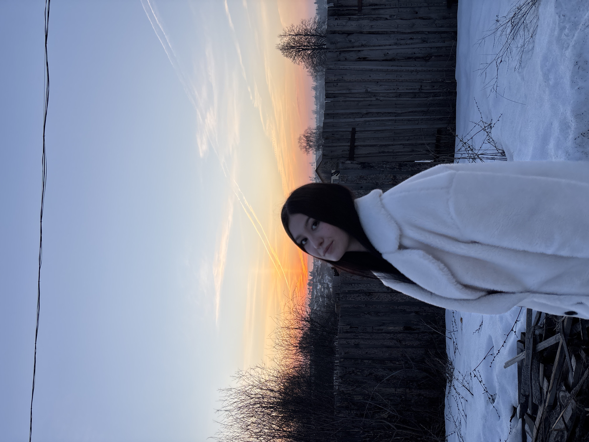 The image captures a young woman standing outdoors in a snow-covered area with a wooden fence in the background. She is wearing a white, fur-like coat. The sky transitions from blue to orange and yellow, suggesting a sunrise or sunset. Bare trees and branches are visible against the backdrop of the sky. In the distance, there is a hint of buildings, potentially part of the city of Фоки, Russia. A power line is visible in the left corner. The scene appears serene and wintery.