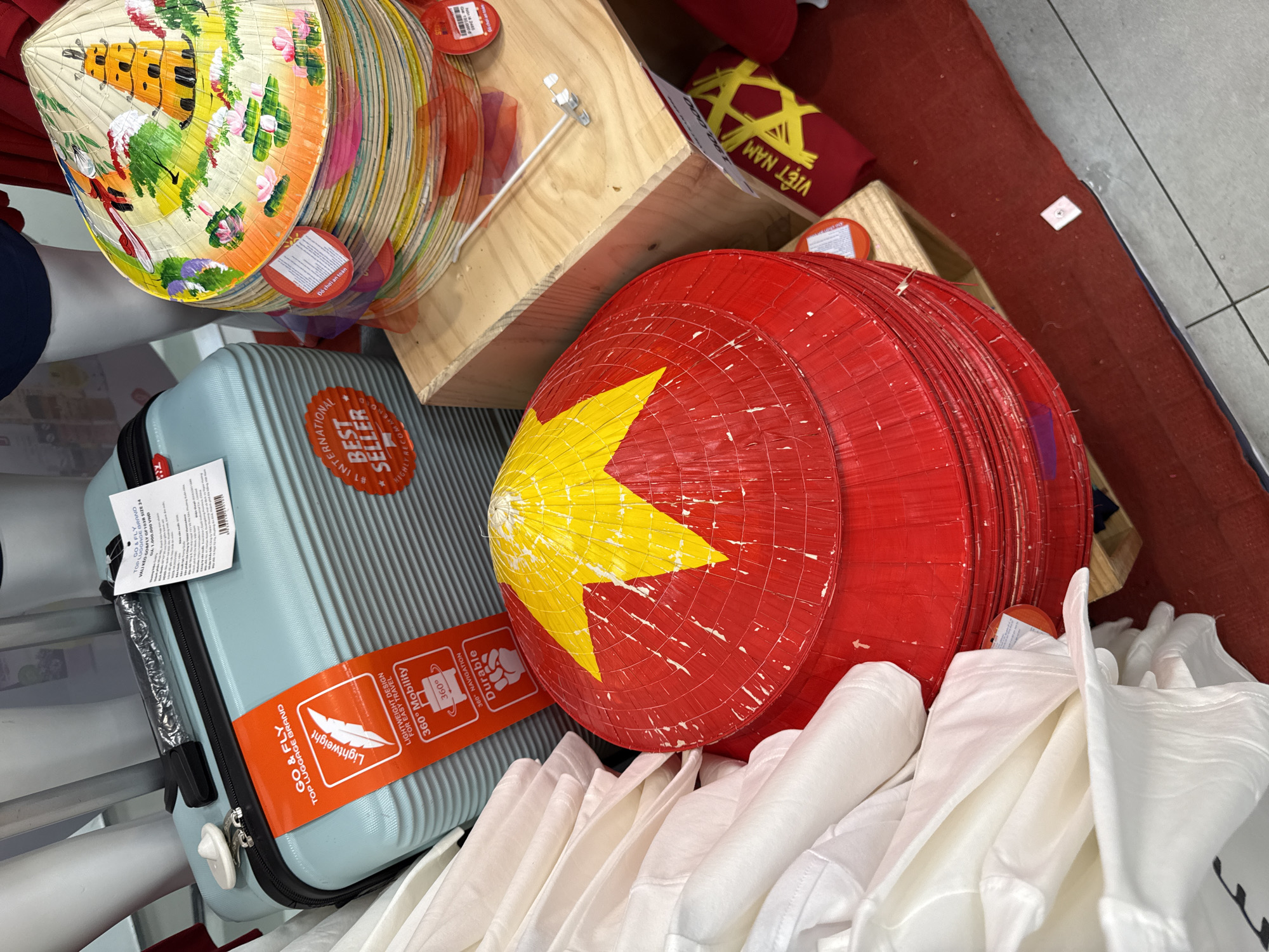 The image shows a close-up, high-angle shot of a display of goods in what appears to be a shop in Nha Trang, Vietnam. In the foreground, a stack of conical hats, known as "nón lá," are prominently displayed. The top hat is red with a yellow star, and the hats below it are multi-colored, featuring painted designs of pagodas and lotus flowers. To the left of the hats, a pale blue suitcase with a "GO & FLY" label is visible. The suitcase has a tag that reads "BEST SELLER." There are also stacks of white garments, possibly t-shirts or fabric, piled on the left side of the frame. The background shows a wooden shelf and more of the conical hats. The overall impression is of a retail setting showcasing traditional Vietnamese handicrafts and travel accessories.