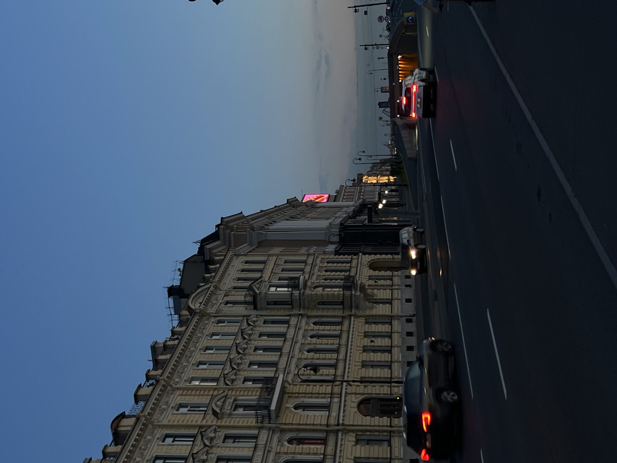 The image shows a street scene at dusk in an urban environment, likely St. Petersburg, Russia, given the architectural style of the buildings. On the left, a grand, ornate yellow building with multiple stories, arched windows, and decorative elements lines the street. A dark-colored SUV with its taillights illuminated is driving in the foreground on the left lane of a multi-lane road. Further down the road, a white car with its brake lights on is approaching an underpass or tunnel. Several streetlights are visible along the road, and the sky is a deep blue, indicating twilight. The road appears to be asphalt, and white lane markings are visible. There are no visible people in the image, but the presence of cars suggests ongoing activity in the city. The overall impression is of a city winding down for the evening, with artificial lights beginning to punctuate the natural light.