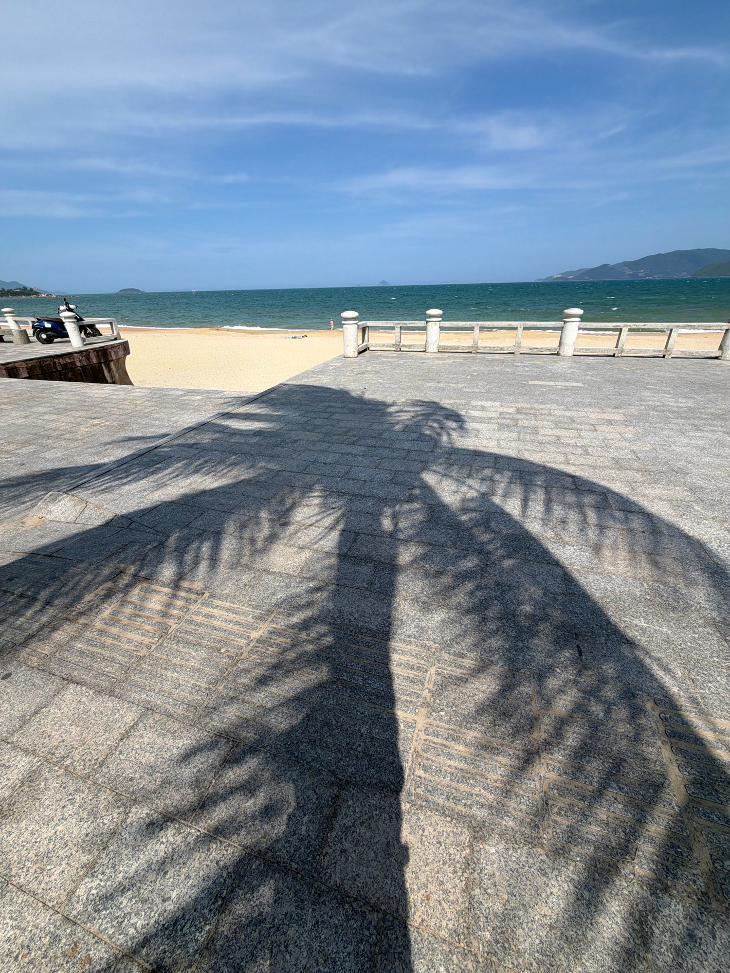 The image depicts a paved walkway overlooking a beach and the ocean. In the foreground, a large shadow of palm fronds stretches across the gray stone pavement. To the left, a dark-colored scooter is parked near a low concrete wall. The walkway leads to a sandy beach, where a few people are visible, one standing in the shallow water and another lying on the sand. A light-colored railing with decorative posts runs along the edge of the walkway overlooking the beach. The ocean is a deep blue-green, and the sky above is clear with scattered white clouds. In the distance, hills or mountains are visible along the coastline. This scene is characteristic of a public area along the coast of Nha Trang, Vietnam, likely a promenade or esplanade designed for public access to the beach.