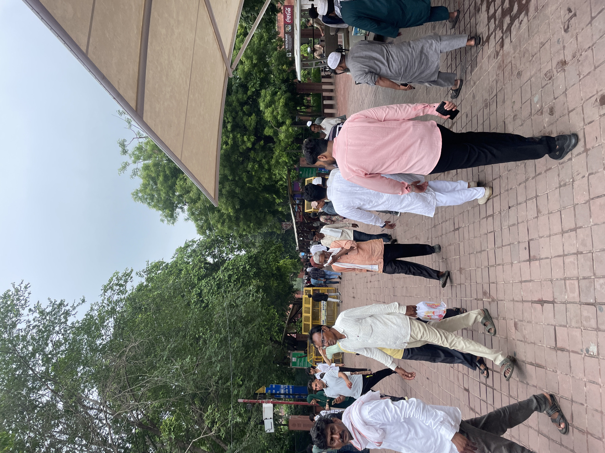 The image depicts a bustling scene on a pedestrian walkway paved with reddish-brown tiles, likely in Agra, India, given the architectural cues and attire of the people. The walkway is lined with lush green trees on the left and right, and a large, light-colored awning extends from the upper right, providing shade. A crowd of people, predominantly men dressed in traditional Indian clothing like kurtas, dhotis, and lungis, are walking in various directions. Some individuals are wearing white and pastel-colored kurtas and trousers, while others are in darker hues.
In the foreground, a man in a white kurta and grey trousers walks towards the viewer, his face partially obscured by a white cloth wrapped around his neck. To his left, another man in a white kurta and dark trousers walks away, his back to the camera. Further back, a group of men are engaged in conversation or walking together. In the midground, yellow metal barricades are visible, suggesting a restricted or designated area, possibly for security or crowd control. Behind the barricades, a white cart with an awning and a Coca-Cola advertisement is parked, and a person in a uniform appears to be attending to it. Further in the distance, more people are walking, and there are glimpses of buildings and foliage. The overall atmosphere is that of a busy public space, likely a tourist attraction or a marketplace, with moderate foot traffic under an overcast sky.