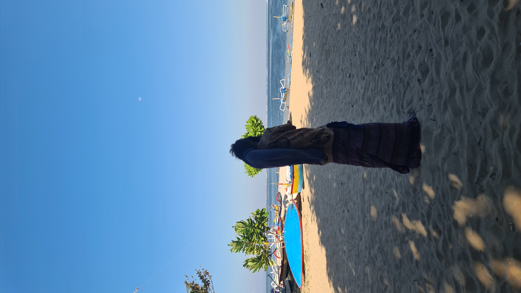 A person stands facing away from the camera on a sandy beach in Denpasar, Indonesia. The person is wearing a dark, patterned skirt and a patterned top, with a blue cloth draped over their shoulders. The beach is lined with several colorful traditional Balinese boats, some resting on the sand and others partially in the shallow water. Two trees with green foliage are visible behind the person. The ocean stretches to the horizon under a clear blue sky, with a faint crescent moon visible. The foreground is dominated by sand with dappled shadows cast by unseen trees.