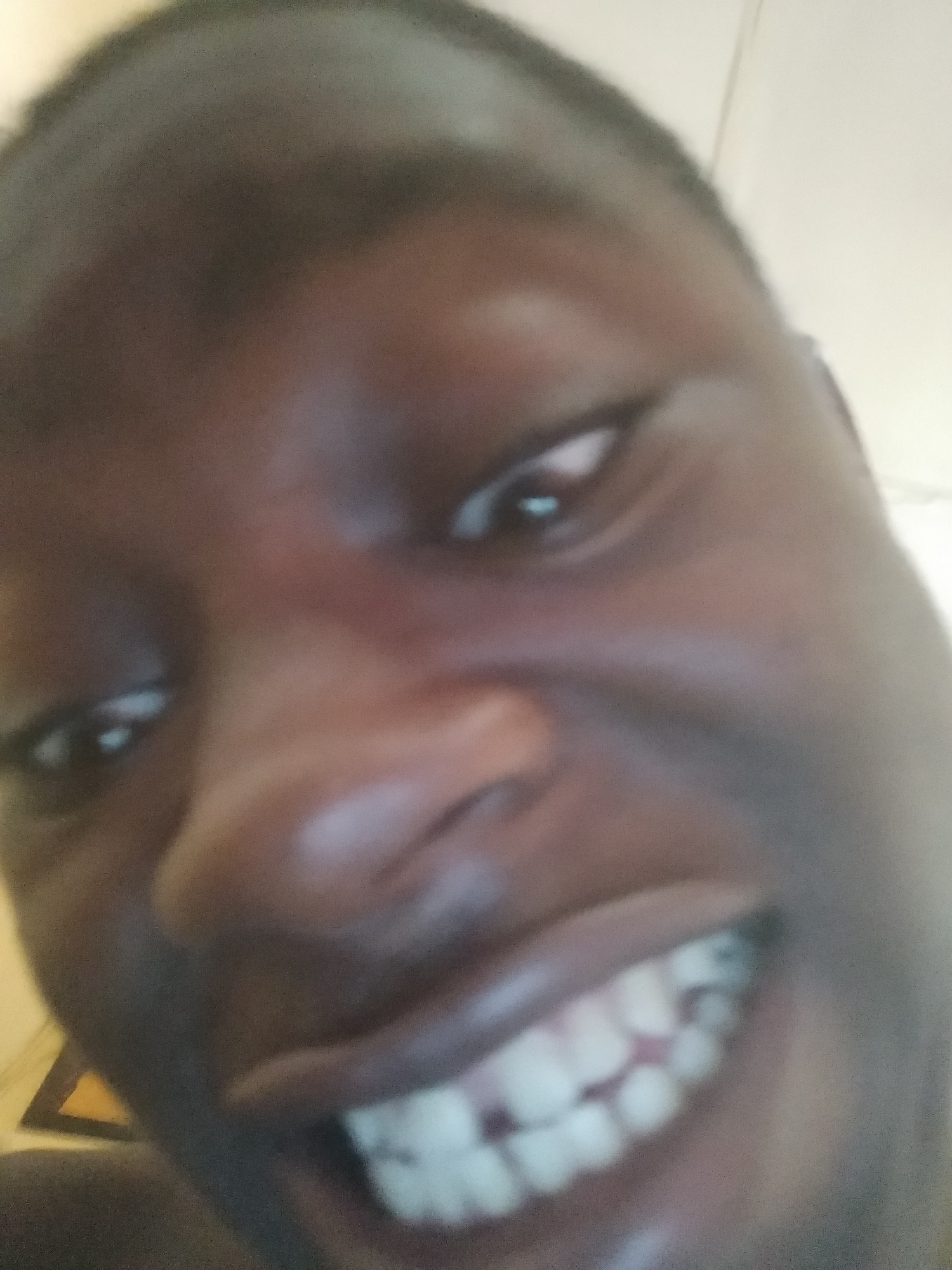 A close-up selfie of a dark-skinned man smiling with his teeth showing. The location, Shangisha, Nigeria is not evident in the image.