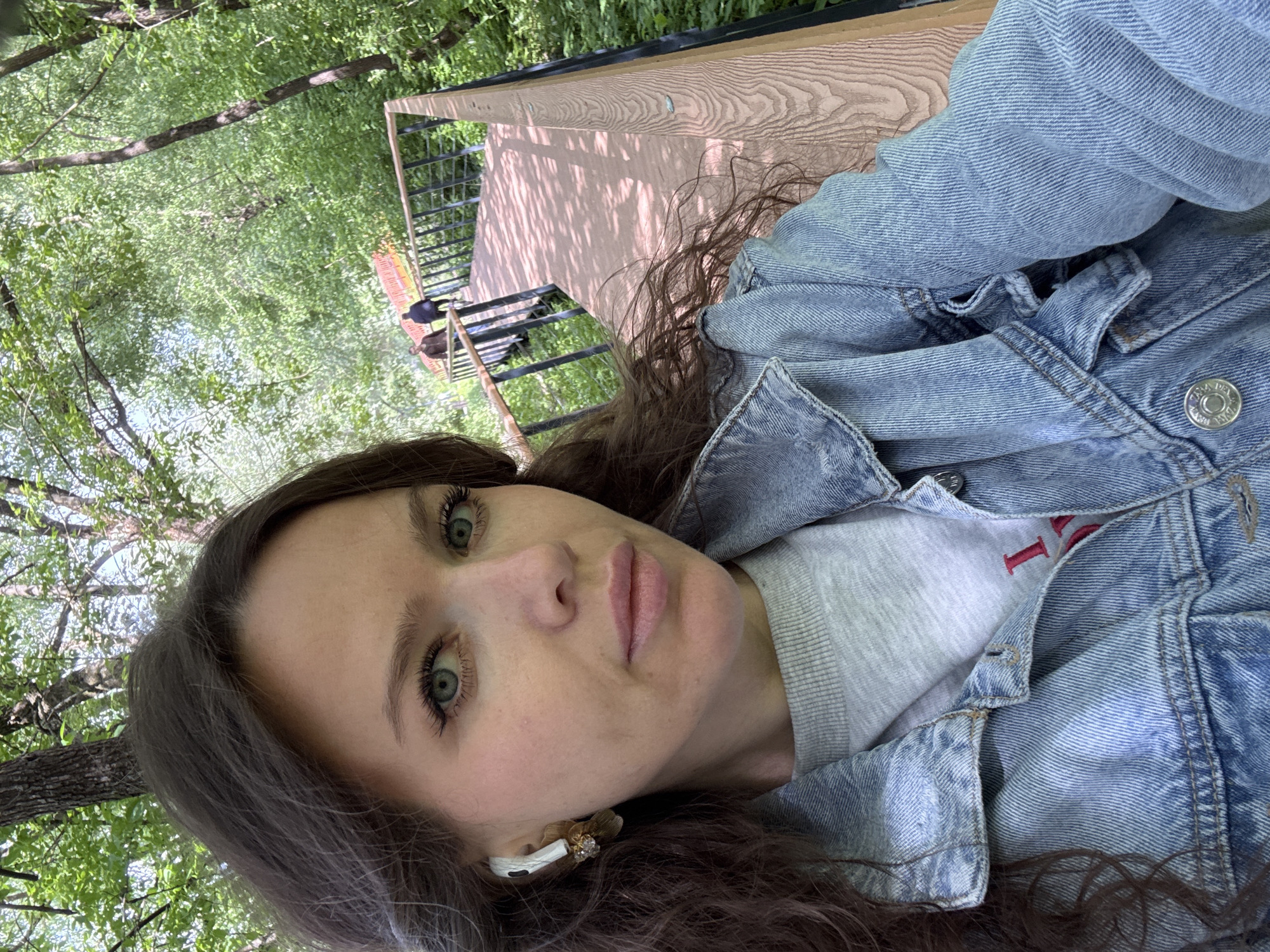 A woman with long, wavy brown hair and green eyes is taking a selfie. She is wearing a light blue denim jacket over a gray sweatshirt. She has a white earbud in her left ear with a floral embellishment. The background shows greenery, suggesting a park or wooded area. There is a wooden platform with railings visible behind her, indicating a raised walkway or bridge. Based on the architecture and environment, it is plausible that this could be a park or recreational area in Moscow, Russia.