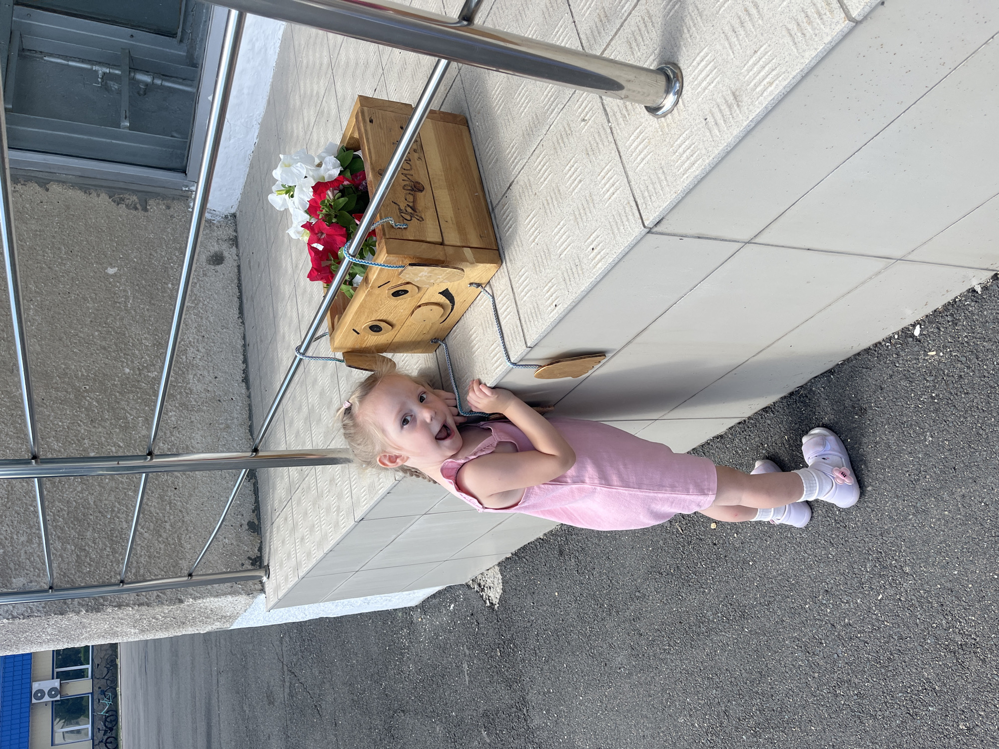 A young girl with blonde pigtails, wearing a pink dress and white socks with light pink shoes adorned with flower details, is captured in an outdoor shot. She is positioned on a grey tiled surface next to a cobblestone pathway. The girl is looking towards the camera with her mouth open in a surprised or excited expression. Behind her, a wooden planter box filled with red and white petunias sits on a metal railing. The planter itself is decorated with a smiley face design made from wooden cutouts. The scene appears to be set during the daytime, with bright sunlight casting shadows, suggesting fair weather. In the background, a building with blue roofing and windows is visible, with some bicycles parked outside. The overall atmosphere is cheerful and highlights the playful interaction of the child with her surroundings.