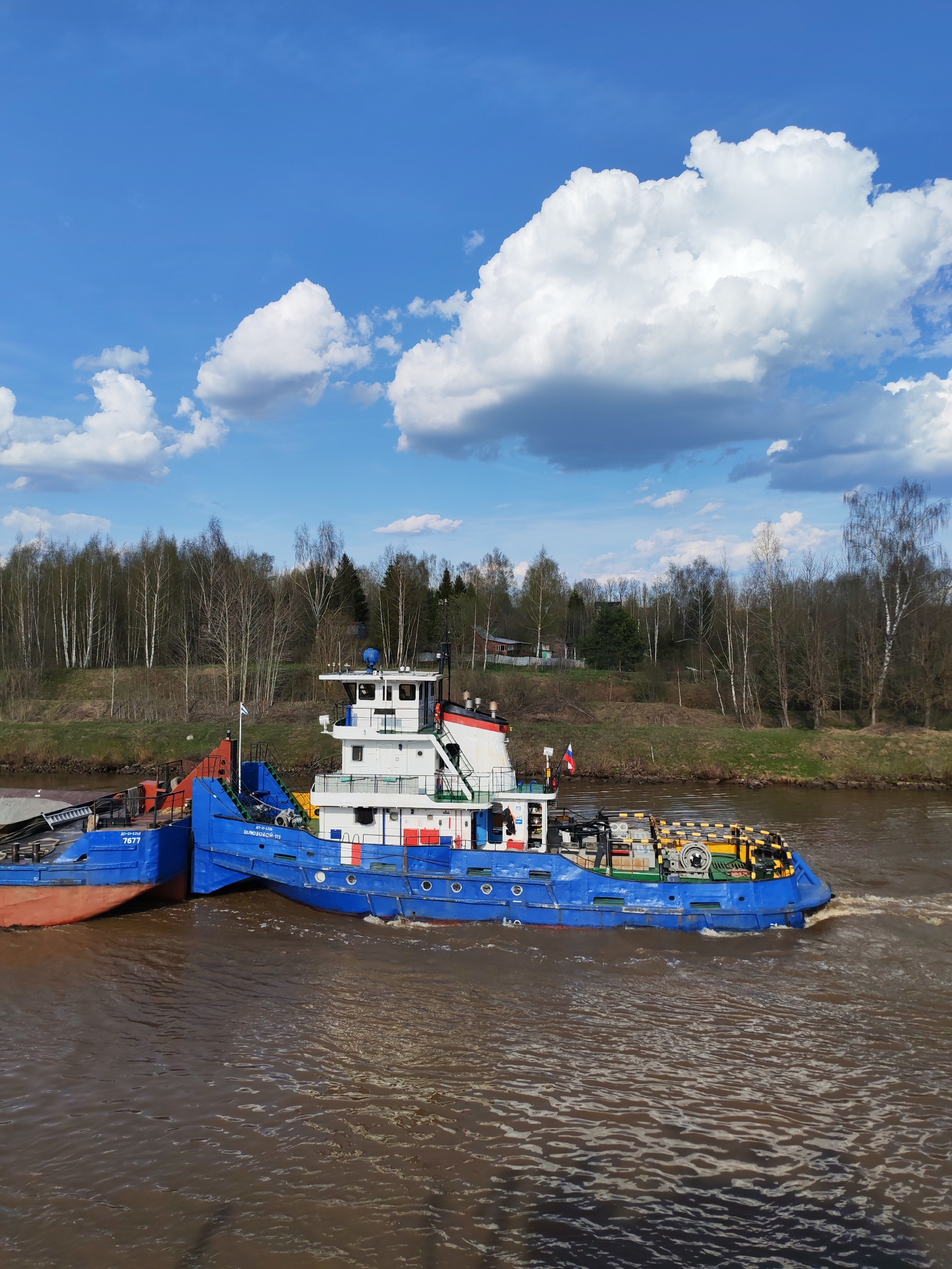 A blue and white tugboat moves along a muddy river in Yakhroma, Russia, on a bright, cloudy day. The boat is connected to a barge. Trees line the riverbank behind the boat.