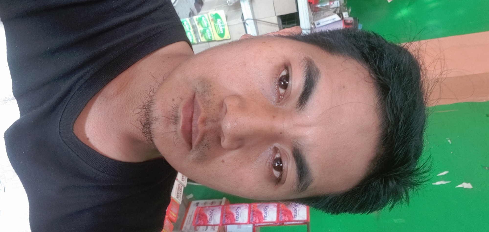 A medium close-up, eye-level, selfie-style photograph shows a man's face and upper torso. He is positioned in the center of the frame, looking directly at the camera. The man has dark hair that is messy and stands up in places, with a prominent hairline. His eyebrows are dark and well-defined. His eyes are brown and appear to be reflecting light. His skin is light brown with visible pores and some minor blemishes. He has a light amount of stubble on his chin and upper lip, forming a small goatee. His lips are closed and appear to be a natural pink color. He is wearing a black t-shirt.
The background is out of focus but appears to be an indoor setting. To the left, there is a shelf with various products, including boxes and bottles, some of which have colorful labels. To the right, there are stacked boxes with red and white packaging that have text and images on them, one of which appears to be an advertisement. The wall behind the man is painted green, with some white smudges on it. A light brown, vertical pillar is visible on the far left of the frame.
Based on external information, the image was taken in the city of Kembang Kerang, Indonesia.