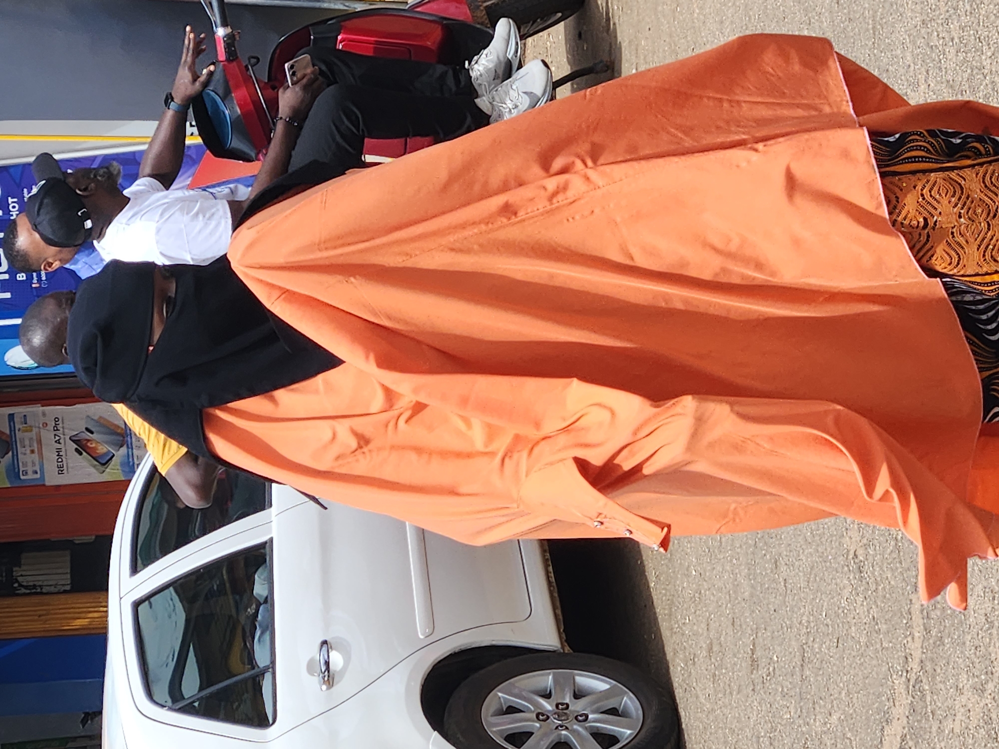 In an outdoor urban setting, a person wearing a black niqab and an orange hijab walks past a white car and a red scooter. The person is in the foreground, obscuring much of the scene. The car is parked on the left side of the image, and its rear wheel is visible. On the right, a red scooter is parked, with two individuals seated on it. The person on the scooter in the back is wearing a black baseball cap, a white t-shirt, and black pants. They are holding a smartphone. The person in front of them is wearing a blue shirt. In the background, a storefront with a yellow pillar and a sign displaying the text "REDMI A7 Pro" is visible. The ground is paved with asphalt.
