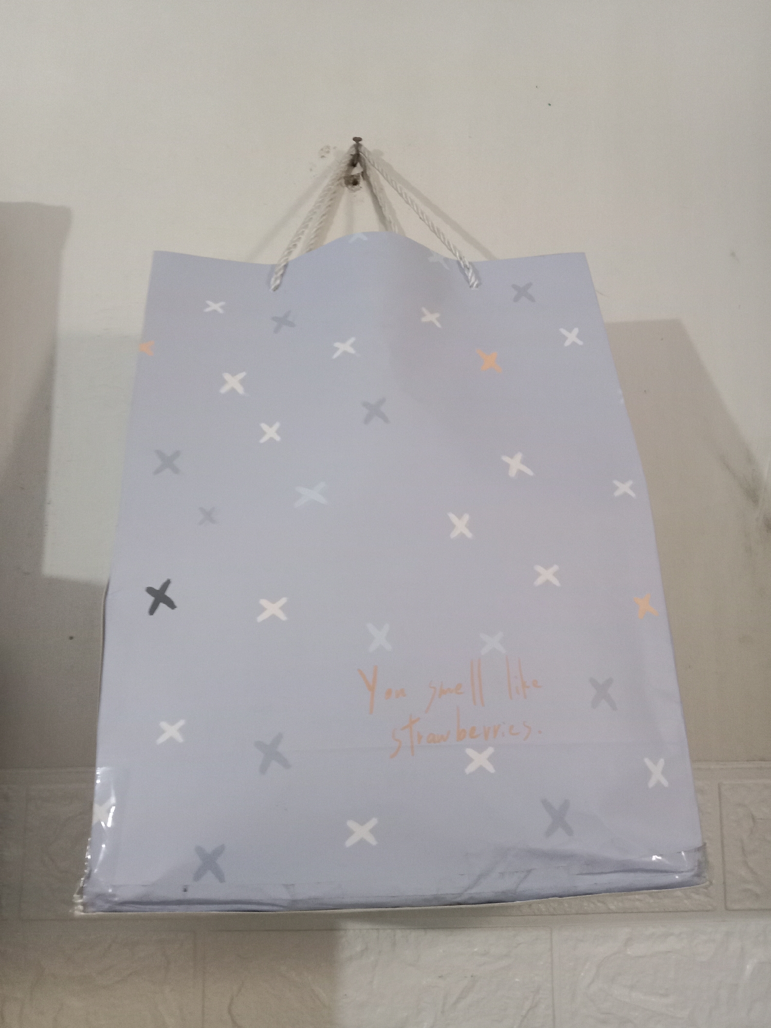 A pale blue gift bag with a white string handle is hanging on a metal hook against a plain white wall. The bag is covered in a pattern of small white, gray, and orange "x" shapes. In the center of the bag, handwritten in an orange script, are the words "You smell like strawberries." The bottom of the bag is covered in clear plastic. The wall has some minor imperfections and shadows. The background below the bag shows a portion of a white tiled surface. This scene is located in the city of Kutosari, Indonesia.