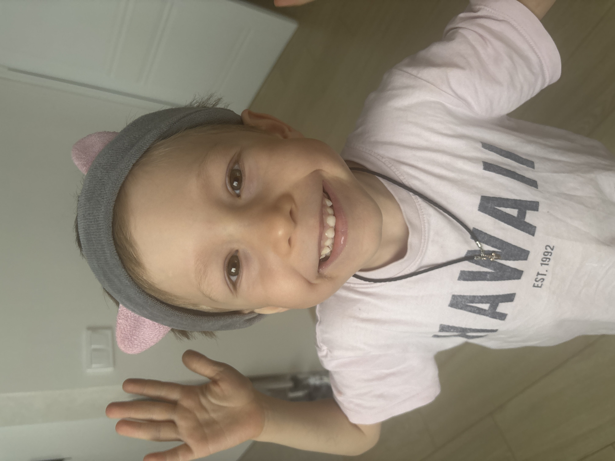 A young child with light skin, light brown hair, and brown eyes is smiling at the camera. The child is wearing a gray headband with pink cat ears and a light pink T-shirt with the word "HAWAII" and "EST. 1992" printed in black. The child is also wearing a black necklace with a silver pendant. The child is holding up their left hand, with the fingers spread apart. The background appears to be a room with white walls and a light-colored floor. The location is Chaikovsky, Russia.